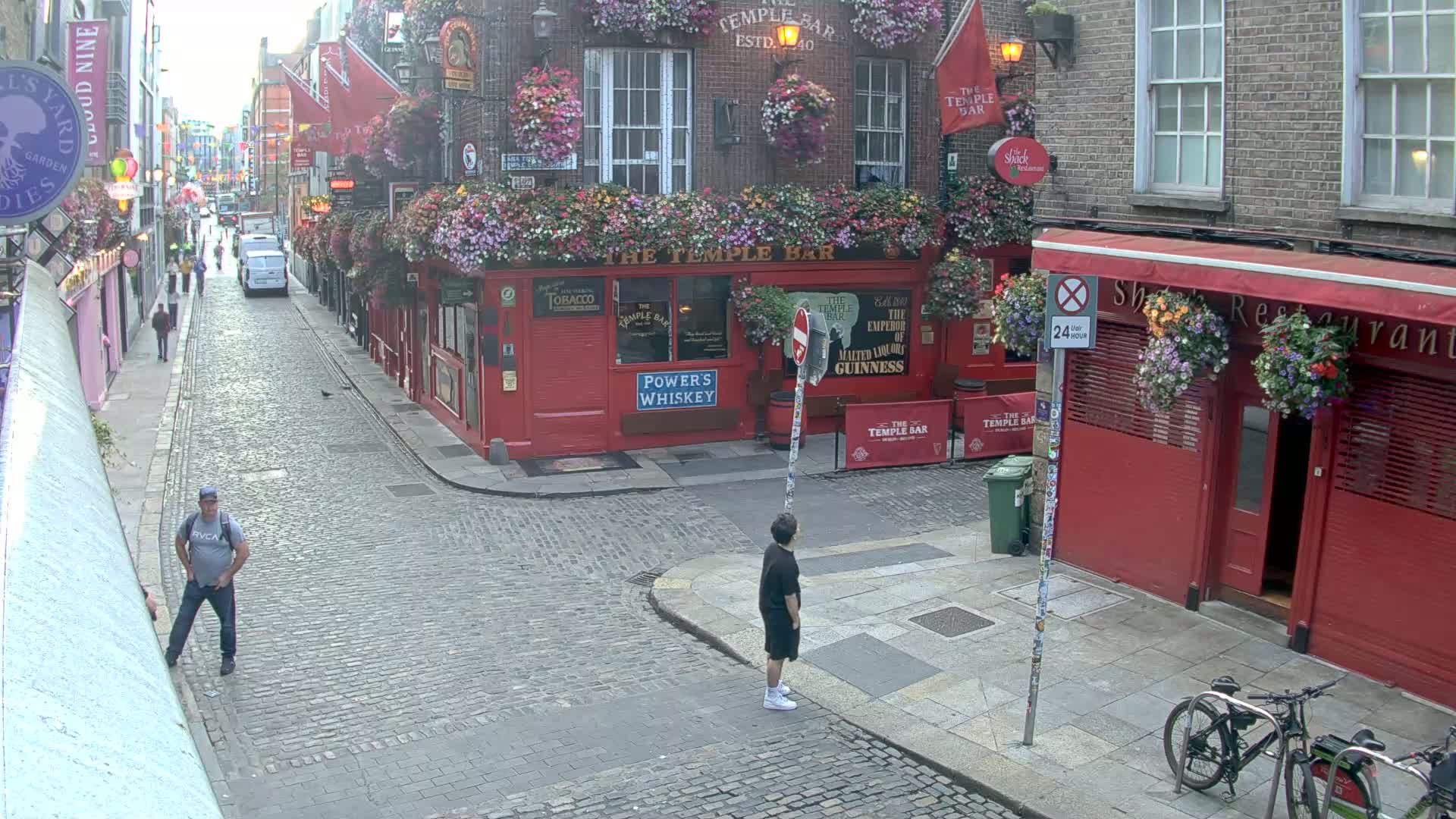 A cobblestone street scene shows two people standing near a red building adorned with hanging flower baskets, with a couple of bicycles parked nearby under overcast skies.