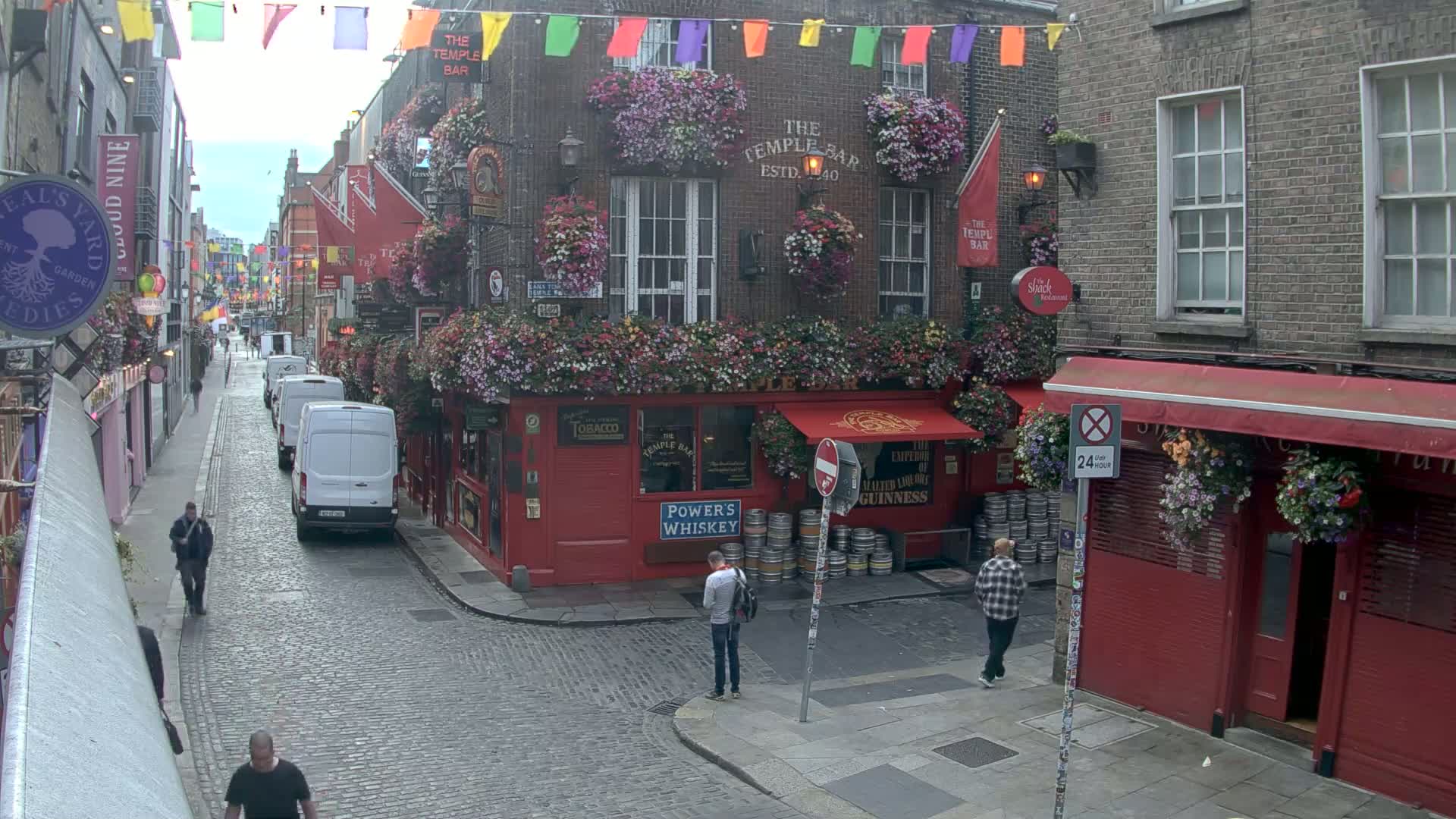 A cobblestone street lined with buildings, many adorned with flower boxes, shows several pedestrians and parked delivery vans on a partly cloudy day.