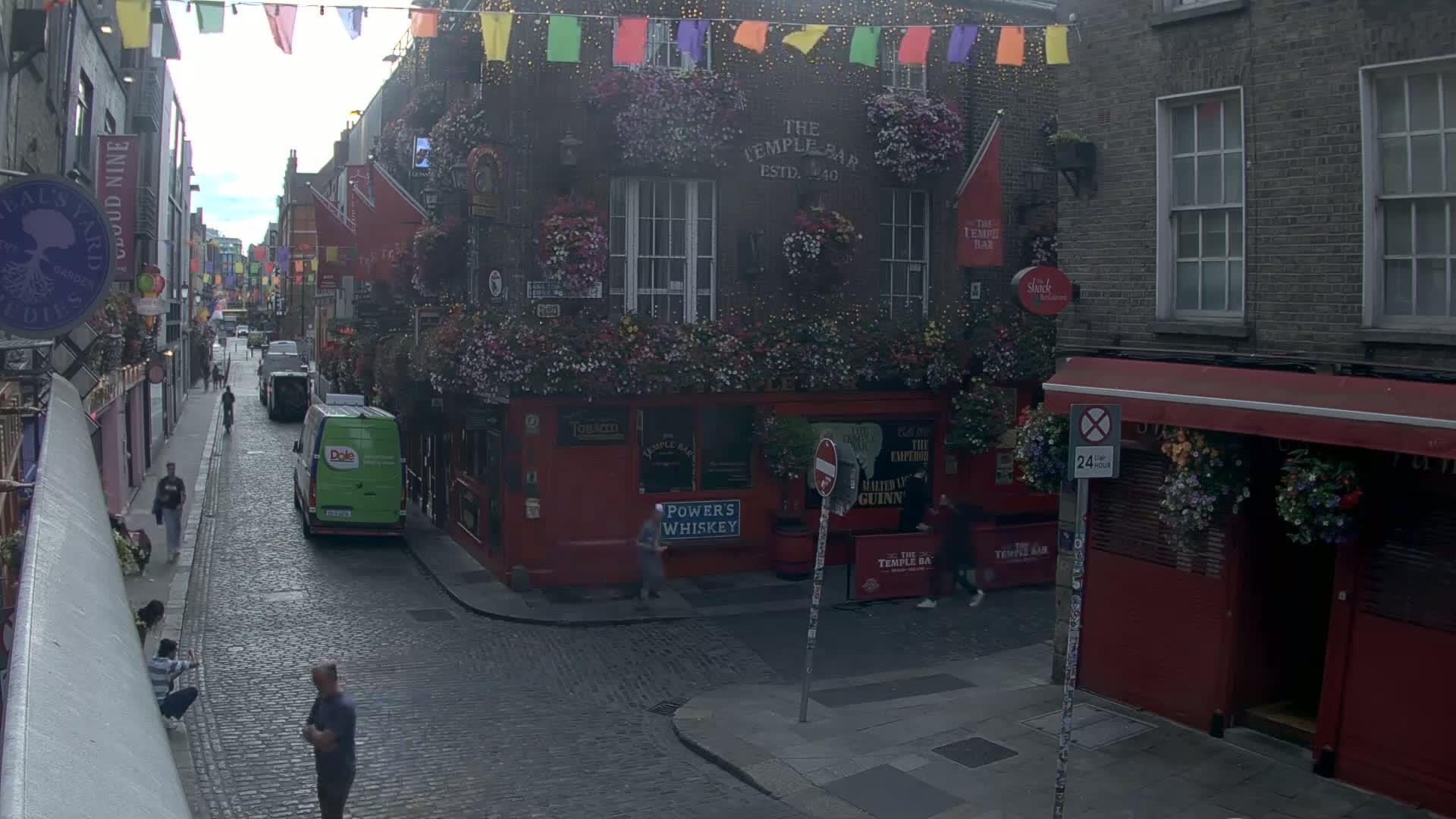A narrow cobblestone street lined with buildings, many adorned with hanging flower baskets, under a partly cloudy sky.