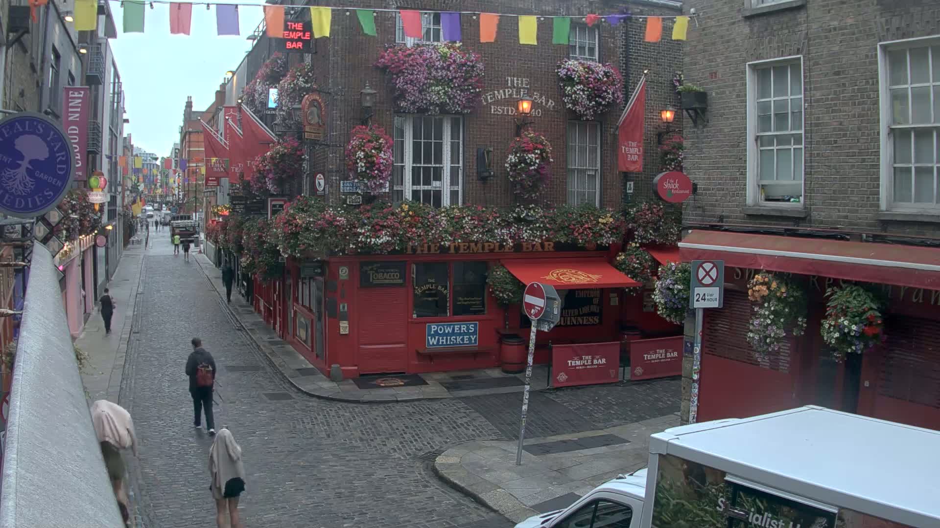 A cobblestone street in a city, overcast, with pedestrians walking past buildings decorated with flower boxes and colorful flags.