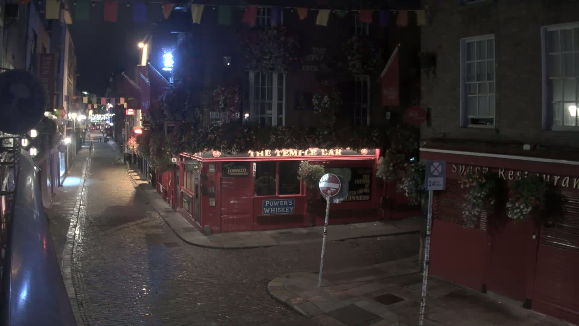 A cobblestone street in a city, overcast, with pedestrians walking past buildings decorated with flower boxes and colorful flags.