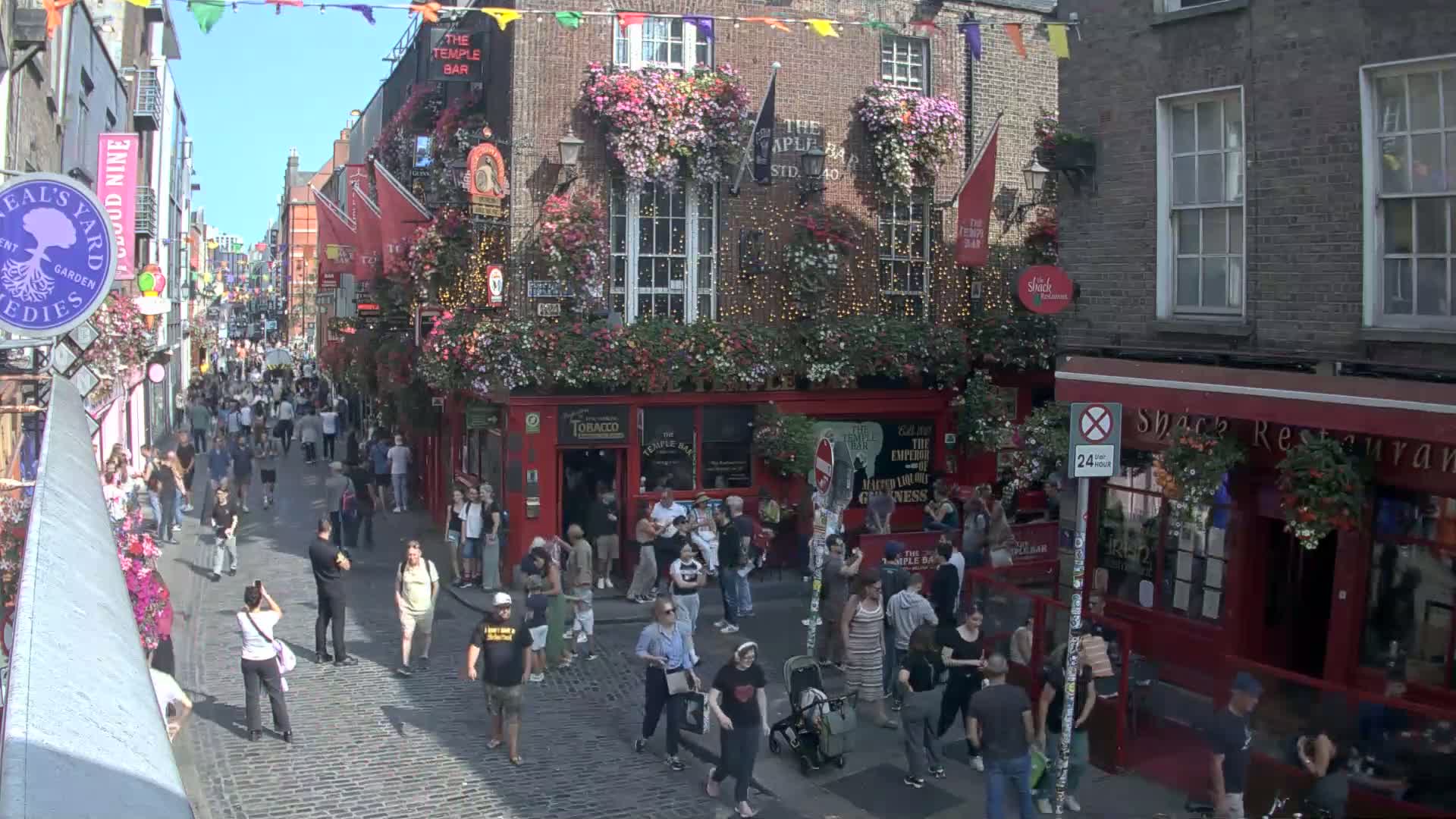 A cobblestone street in a city, overcast, with pedestrians walking past buildings decorated with flower boxes and colorful flags.