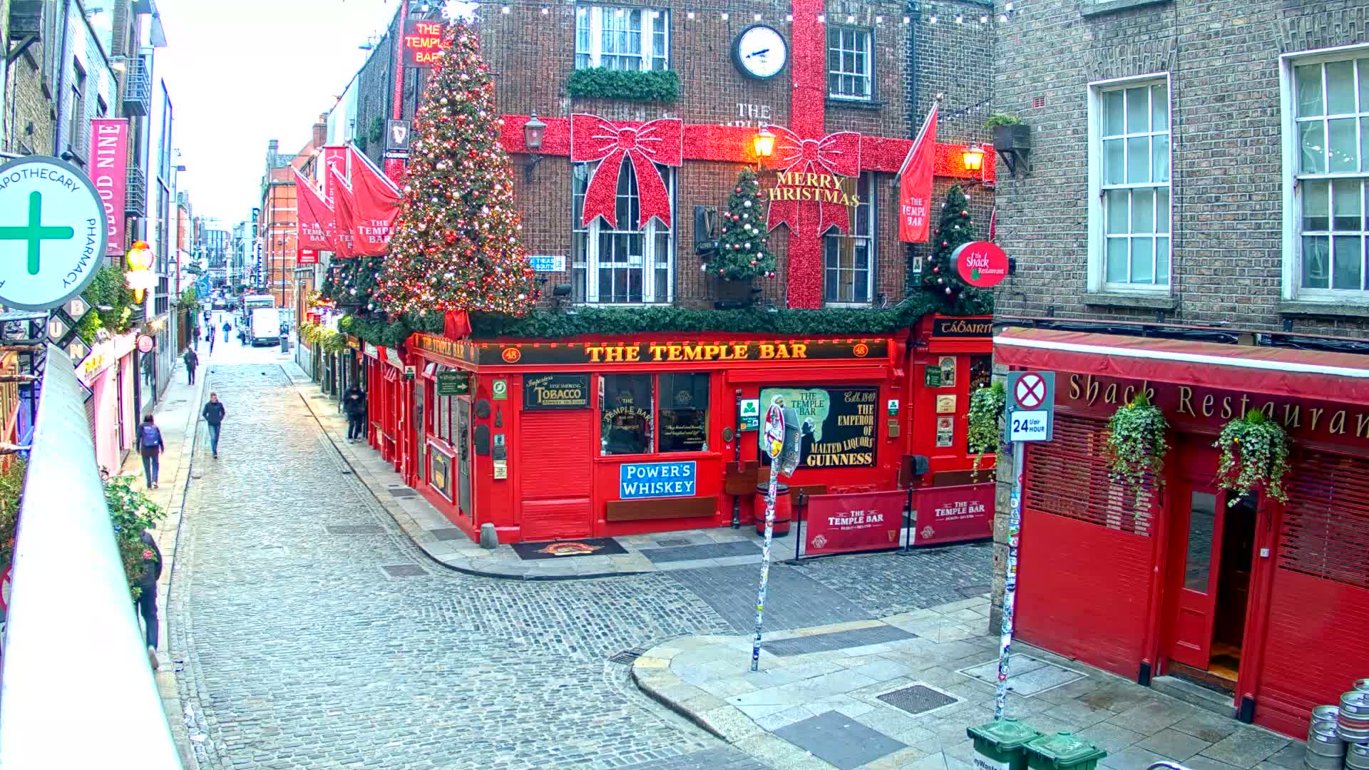 A festive cobblestone street scene shows people walking past a prominent red pub heavily decorated with Christmas lights and bows under an overcast sky.