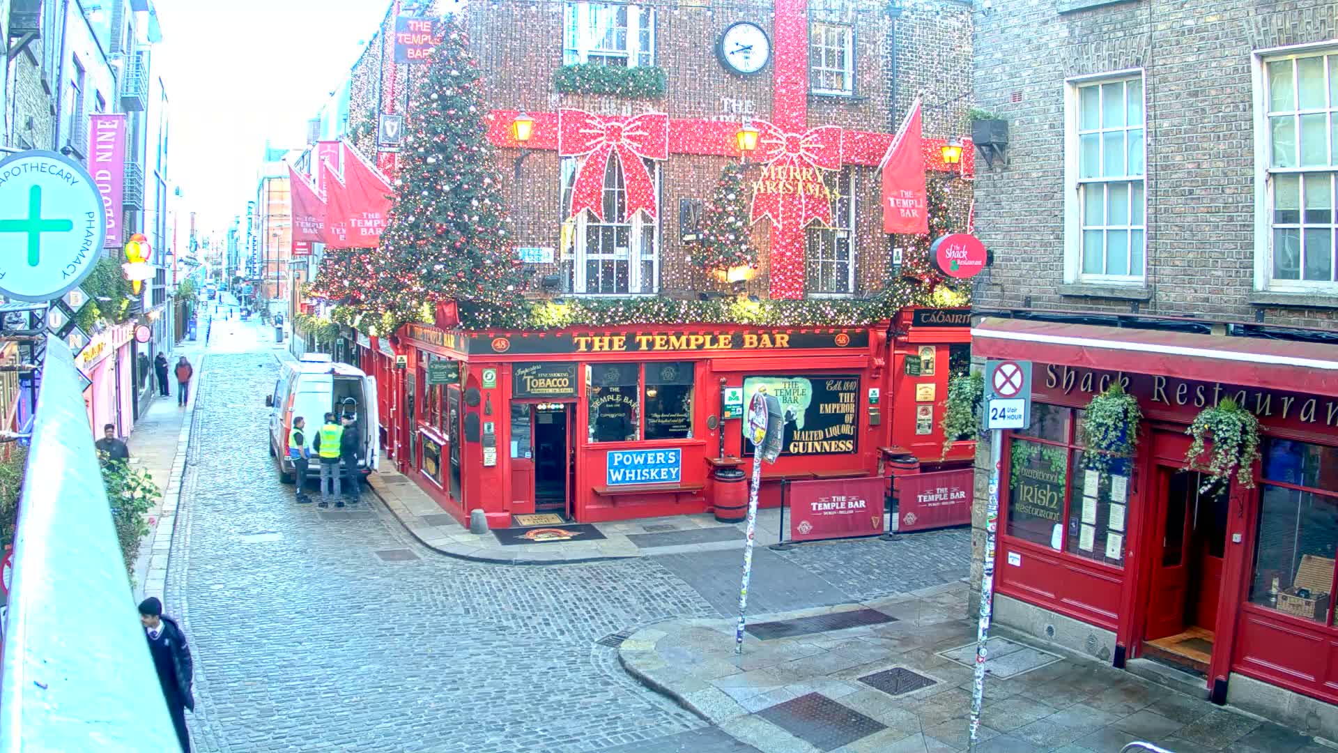 A festive cobblestone street in a European city features buildings extensively decorated for Christmas with red bows, twinkling lights, and evergreen trees, prominently including a large red corner pub, all under bright daylight with several pedestrians and a white van present.