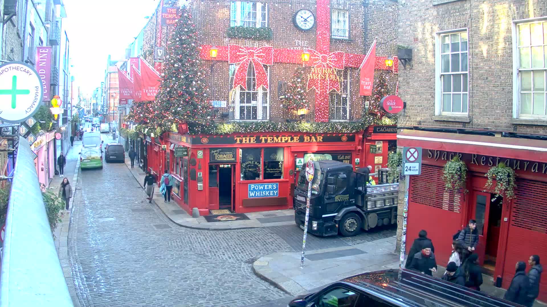 A historic cobblestone street, damp in places, is bustling with pedestrians and vehicles amidst multi-story brick buildings, most notably a bright red corner establishment and others with elaborate Christmas decorations including twinkling lights, large festive bows, and decorated evergreen trees, all under clear, sunny skies.