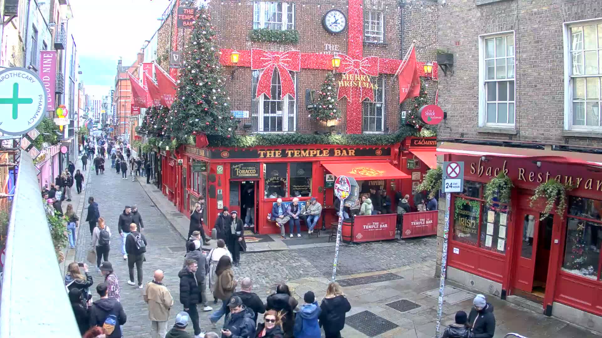 A historic cobblestone street, damp in places, is bustling with pedestrians and vehicles amidst multi-story brick buildings, most notably a bright red corner establishment and others with elaborate Christmas decorations including twinkling lights, large festive bows, and decorated evergreen trees, all under clear, sunny skies.
