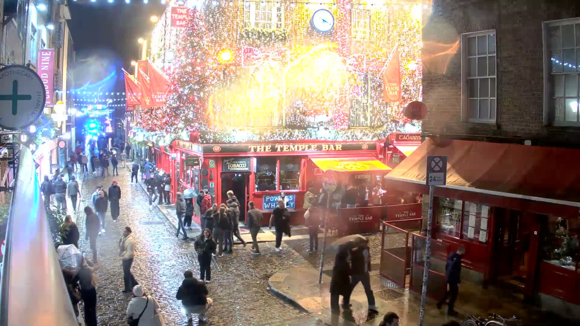 A historic cobblestone street, damp in places, is bustling with pedestrians and vehicles amidst multi-story brick buildings, most notably a bright red corner establishment and others with elaborate Christmas decorations including twinkling lights, large festive bows, and decorated evergreen trees, all under clear, sunny skies.
