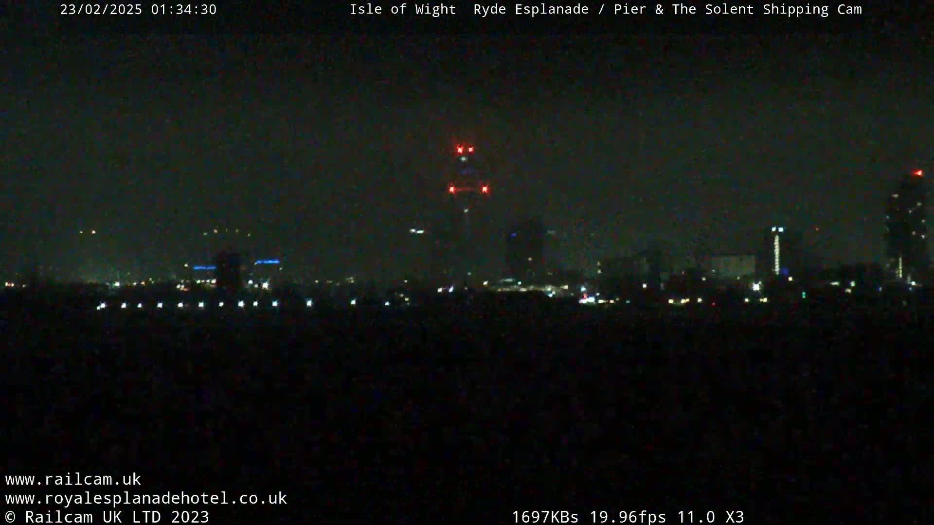 A nighttime long shot shows a dark city skyline with a few illuminated buildings and lights, under a clear, dark sky.