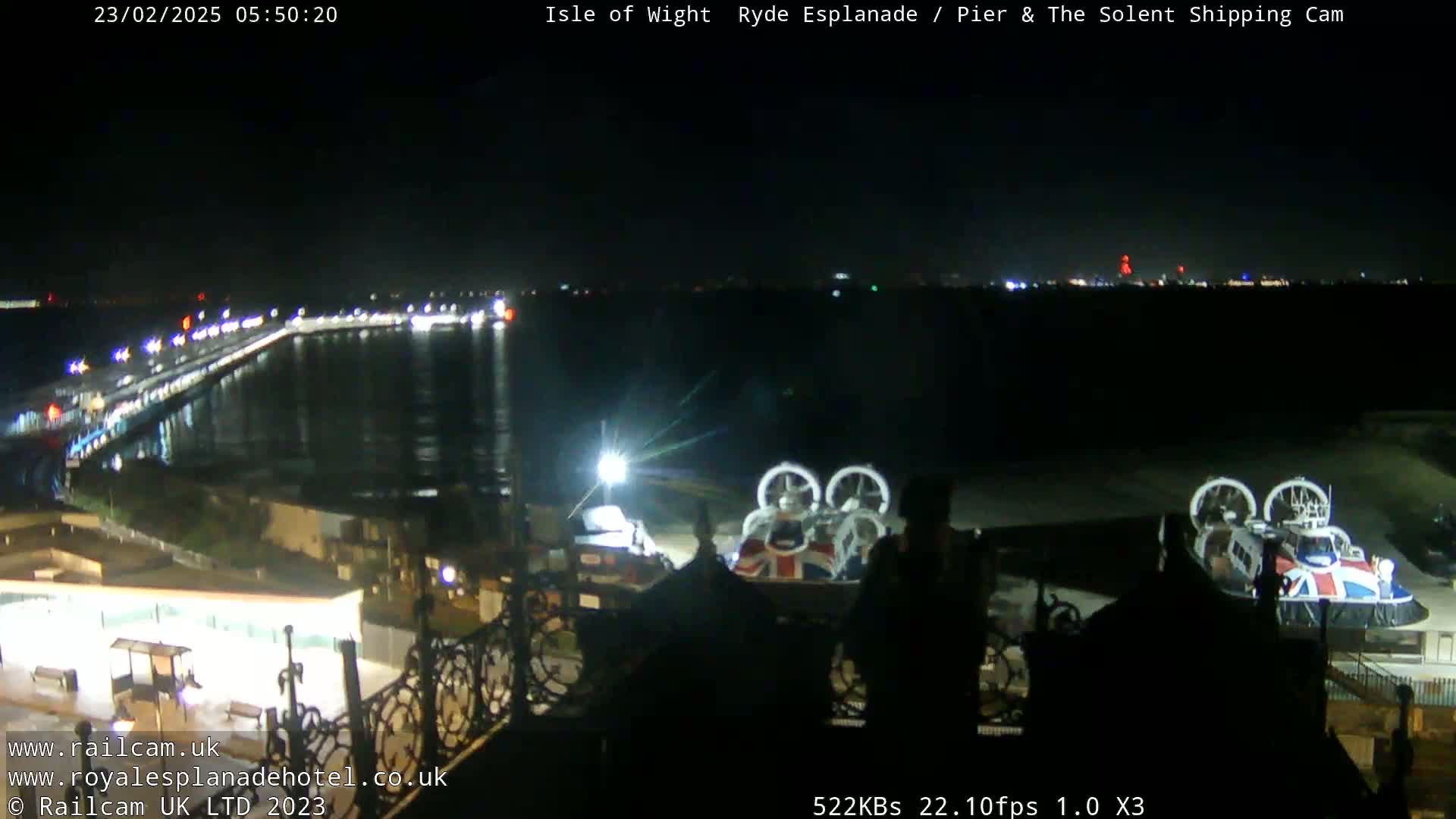 At night, a hovercraft sits on a dock near a lit-up pier and body of water, with a distant city skyline visible.