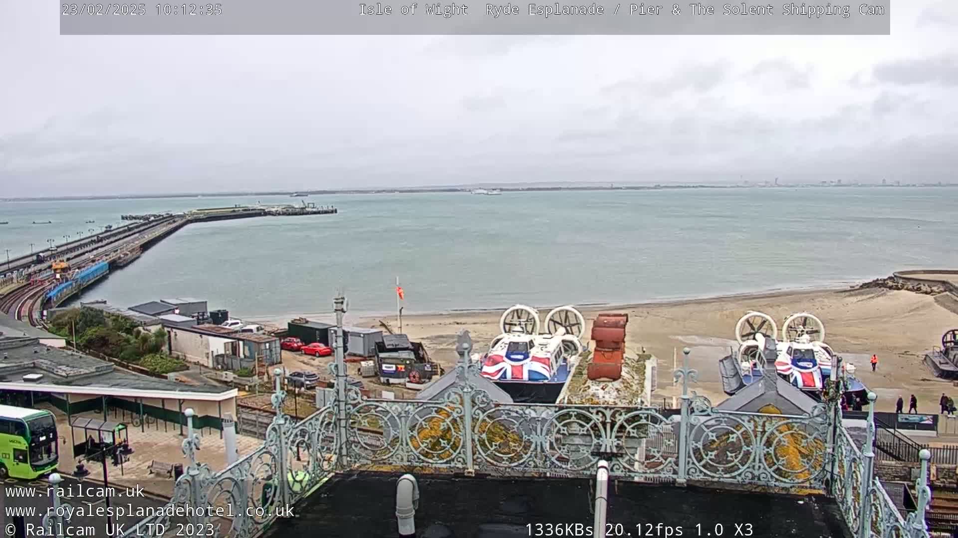 An overcast day reveals a coastal scene with a pier extending into a body of water, a sandy beach with hovercrafts, and a bus visible near buildings.