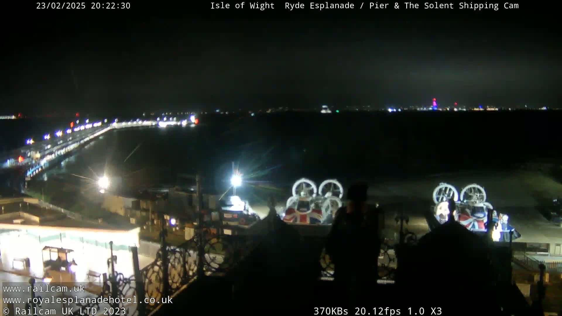 A nighttime view of a pier, a body of water, and a hovercraft, with city lights visible in the distance under a clear, dark sky.