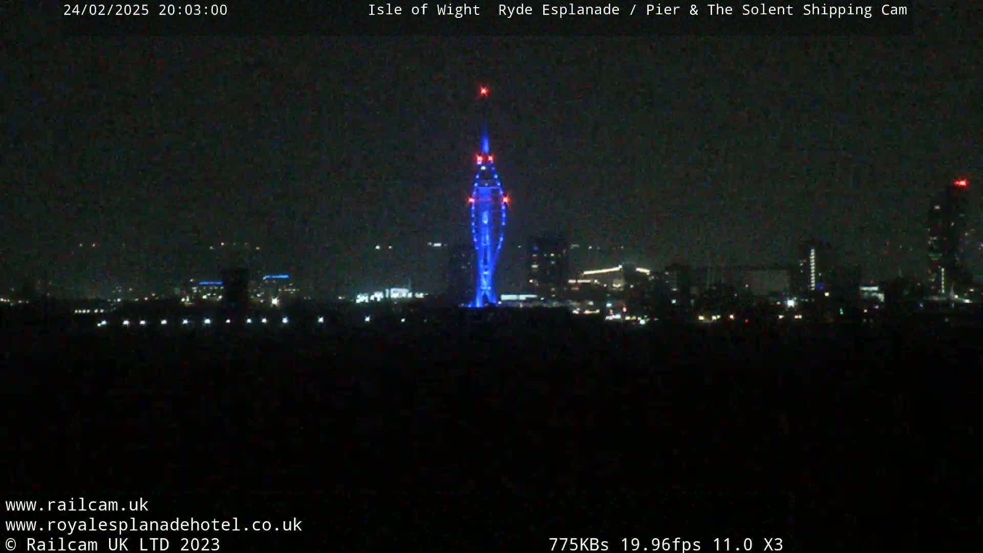 A tall, slender tower, illuminated in blue, stands prominently against a dark, nighttime cityscape.
