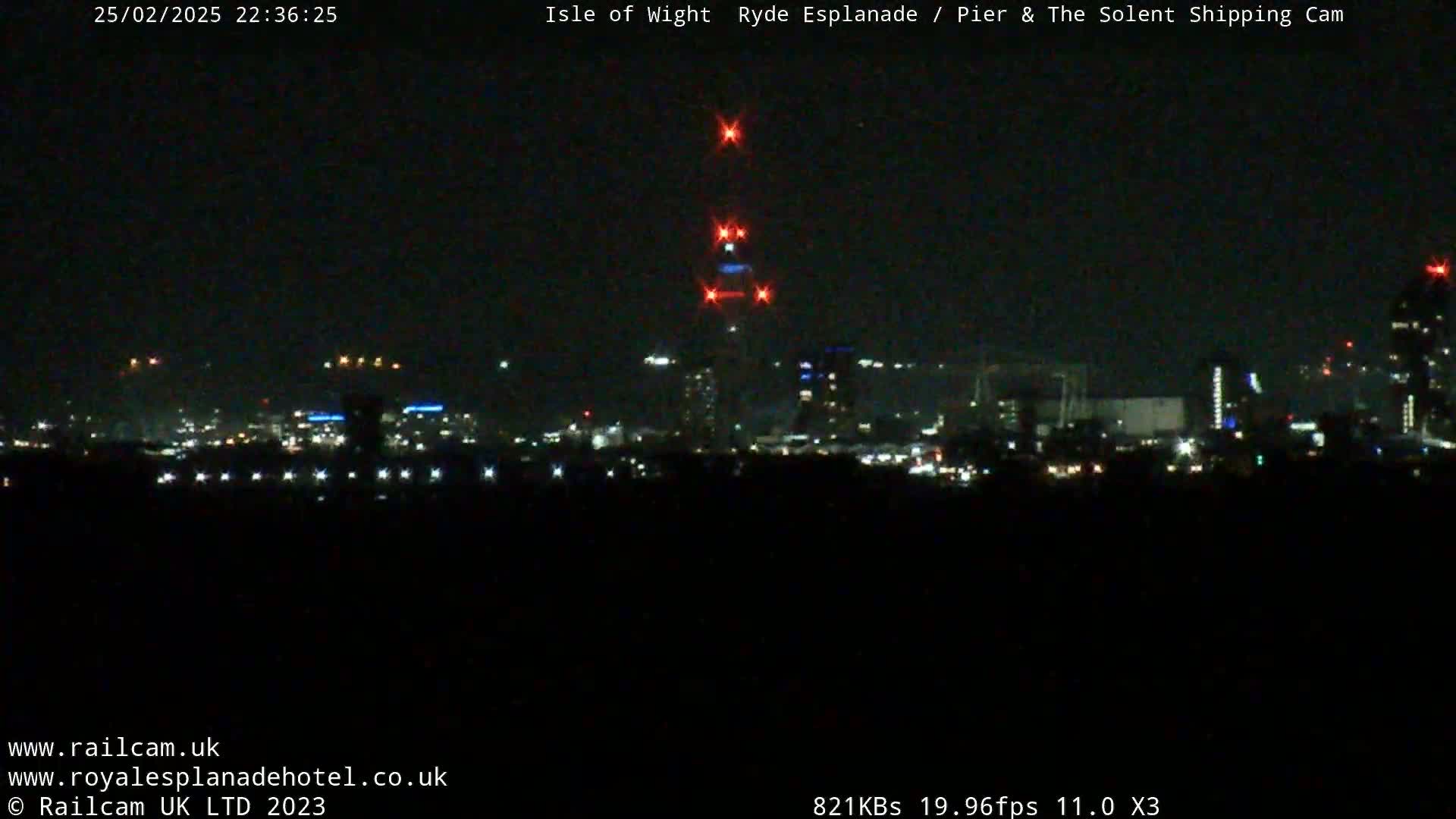 A nighttime long shot of a city skyline illuminated by lights, with a tall structure displaying red lights at its peak, under a clear, dark sky.