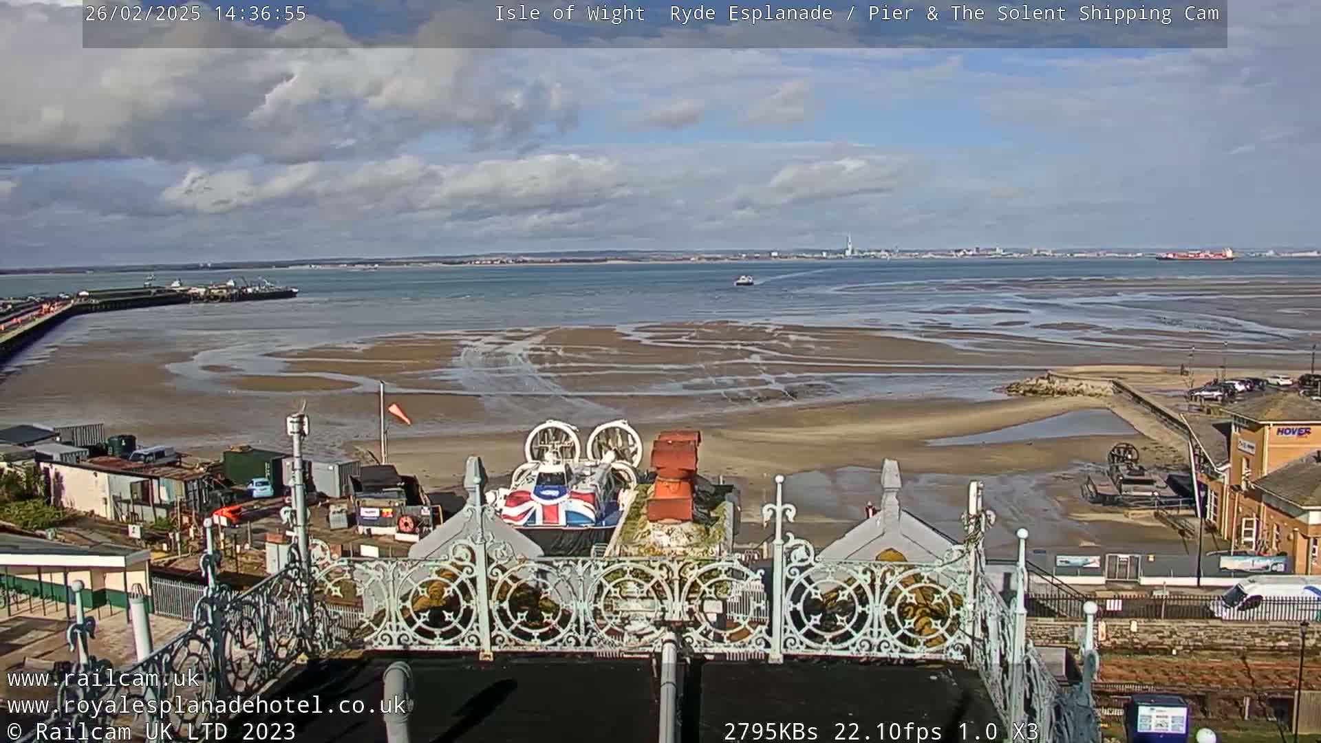 A hovercraft is docked near a pier on a partly sunny day with a partly exposed sandy shore and calm water.