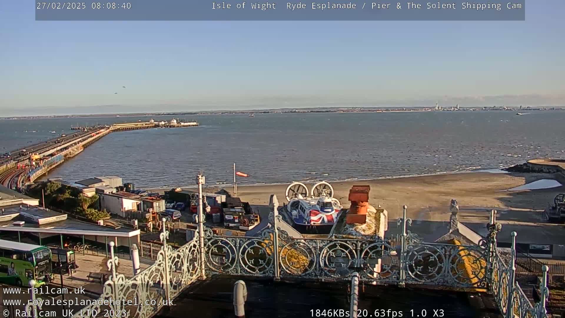 A long pier extends into a calm body of water under a clear, bright sky, with a hovercraft visible on the shore near ornate metal railings.