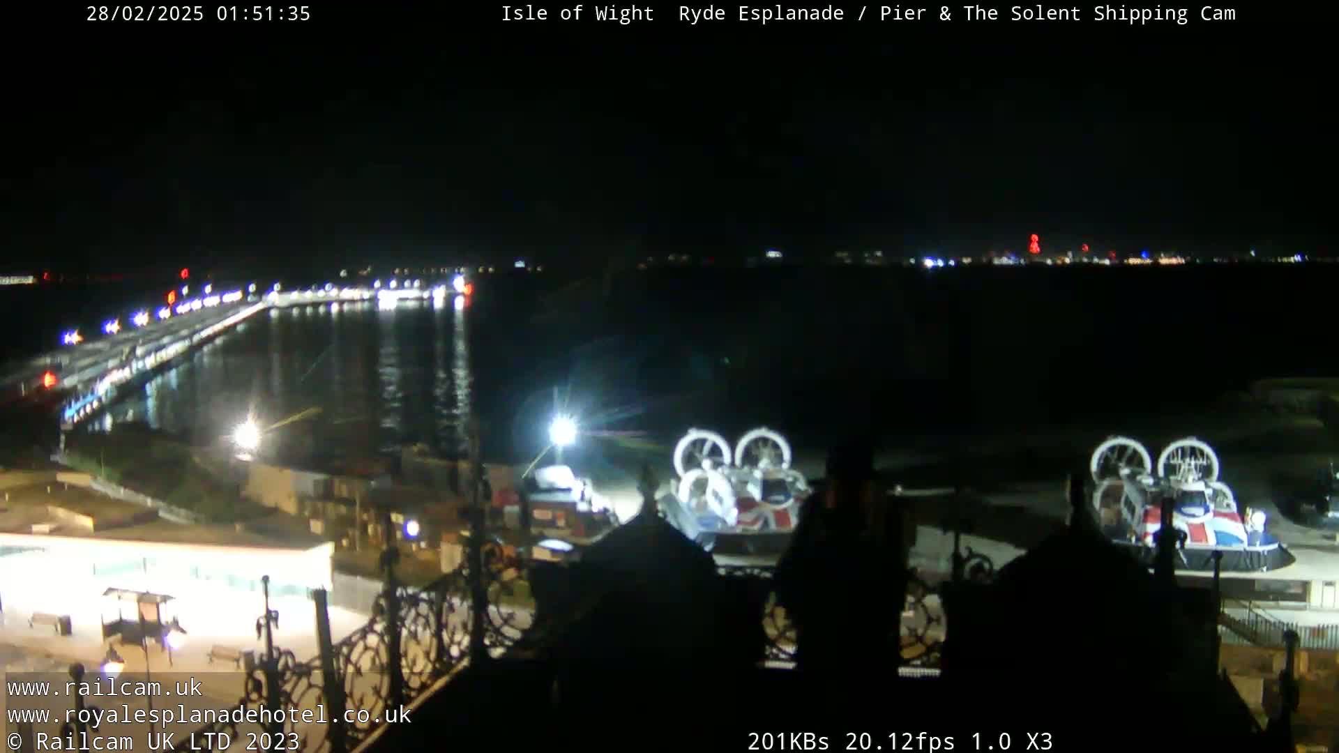A nighttime view of a pier and body of water with lights illuminating a shoreline and a hovercraft parked nearby.