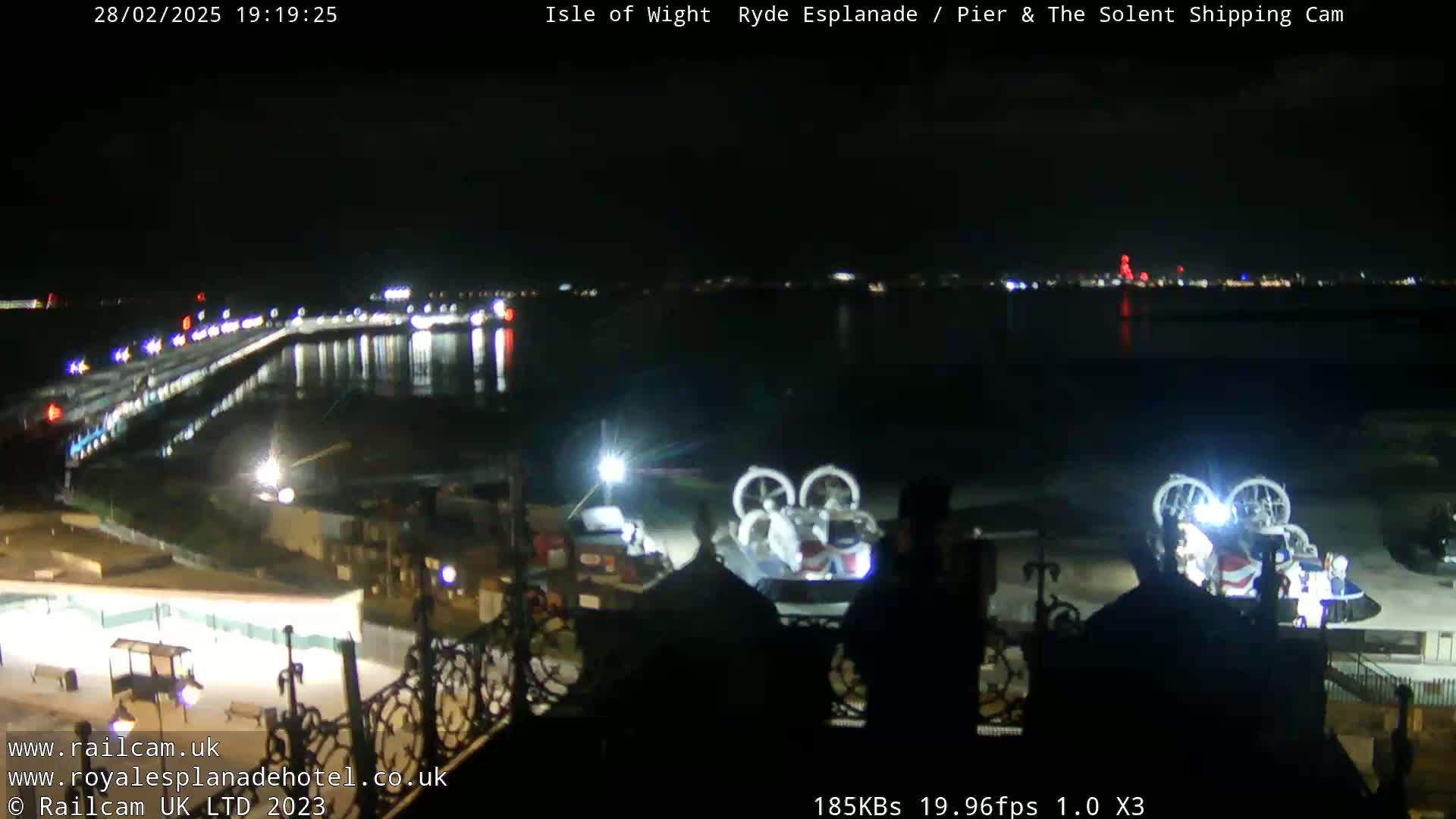 A nighttime view of a body of water with lights reflecting on the surface, a pier extending into the water, and two hovercrafts on land.