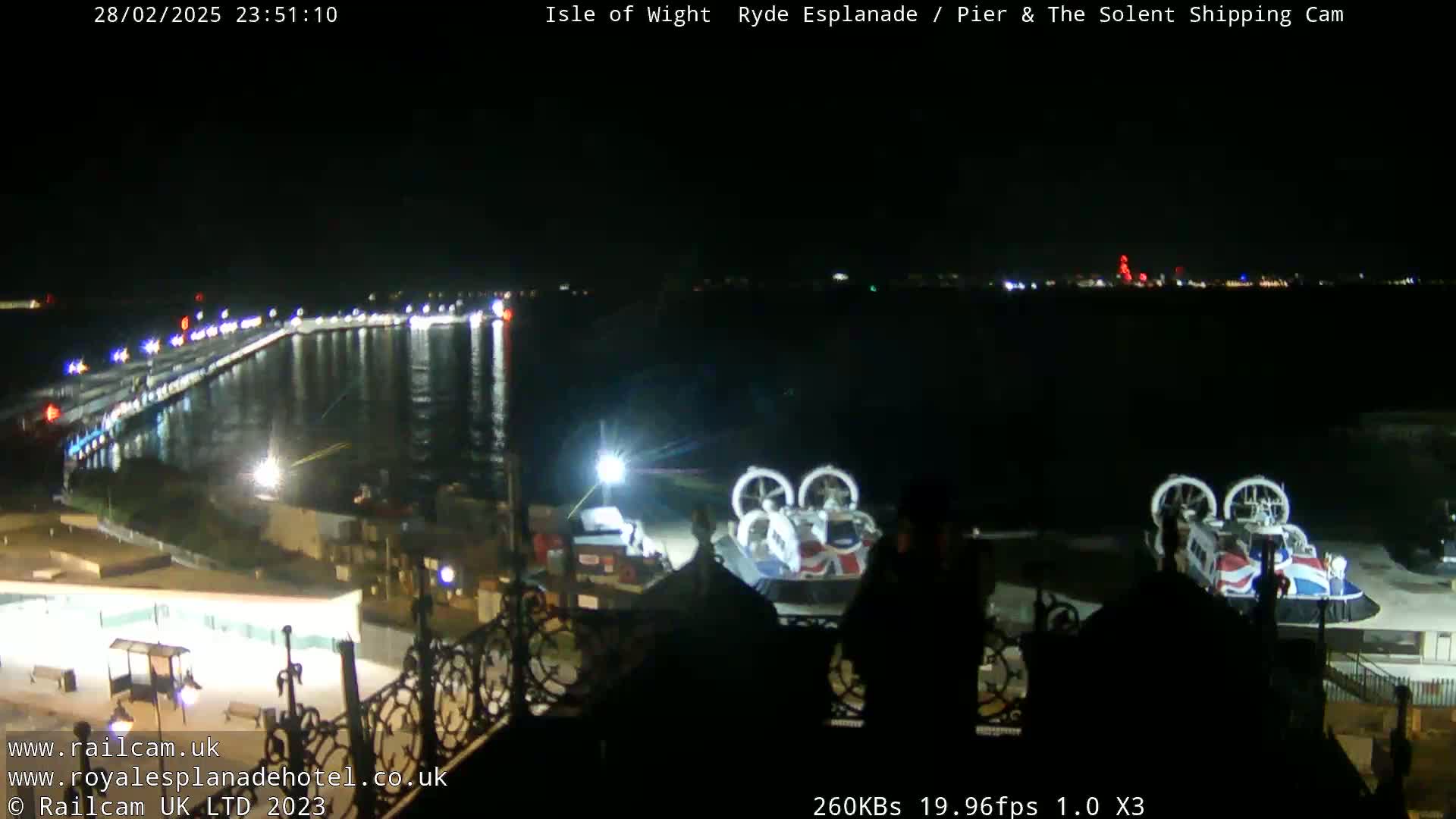 A nighttime view of a harbor with two hovercrafts docked, a lit pier extending into the water, and city lights visible in the distance.