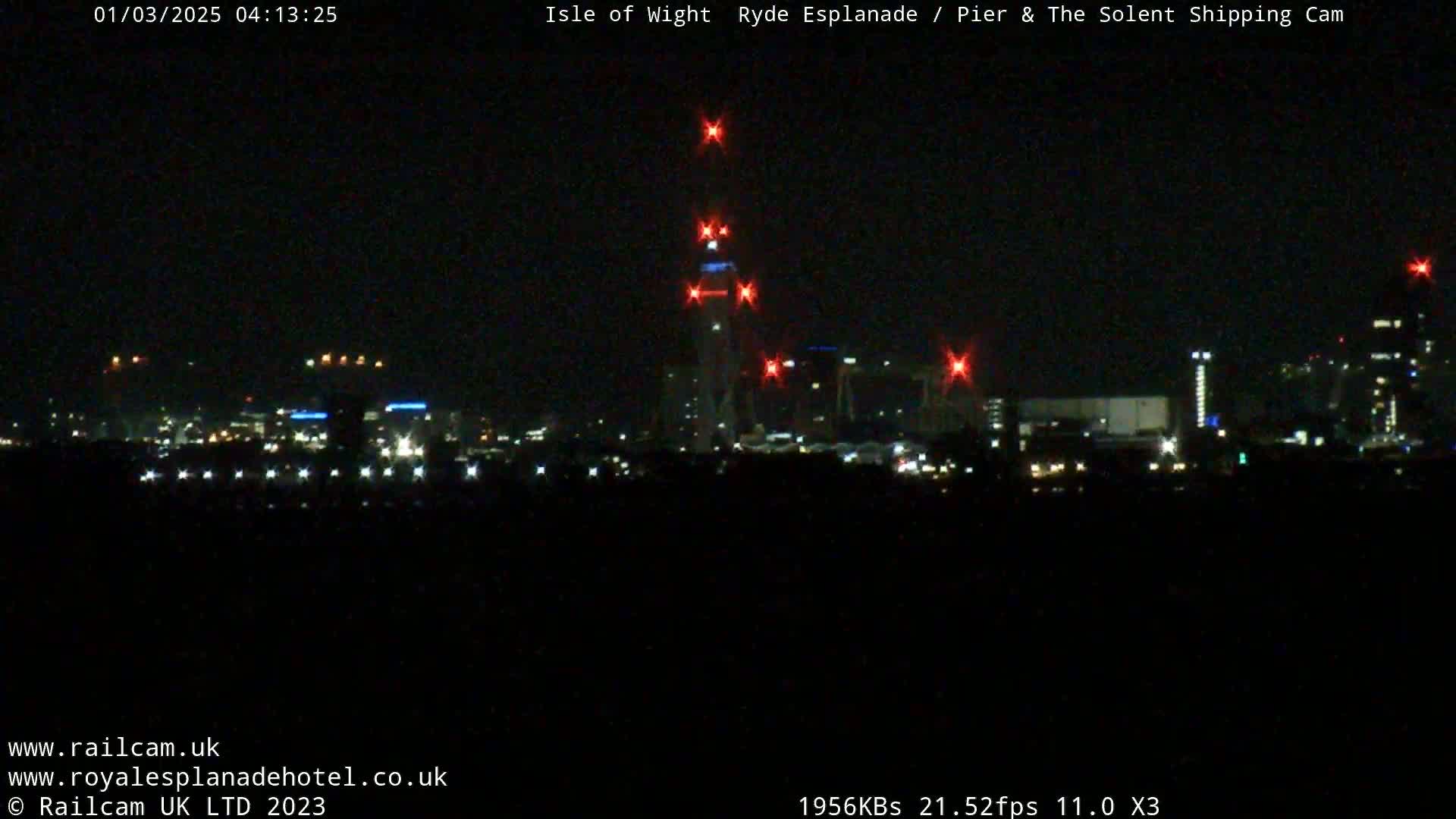 A nighttime long shot of a city skyline with a tall structure illuminated by red lights, under a clear, dark sky.