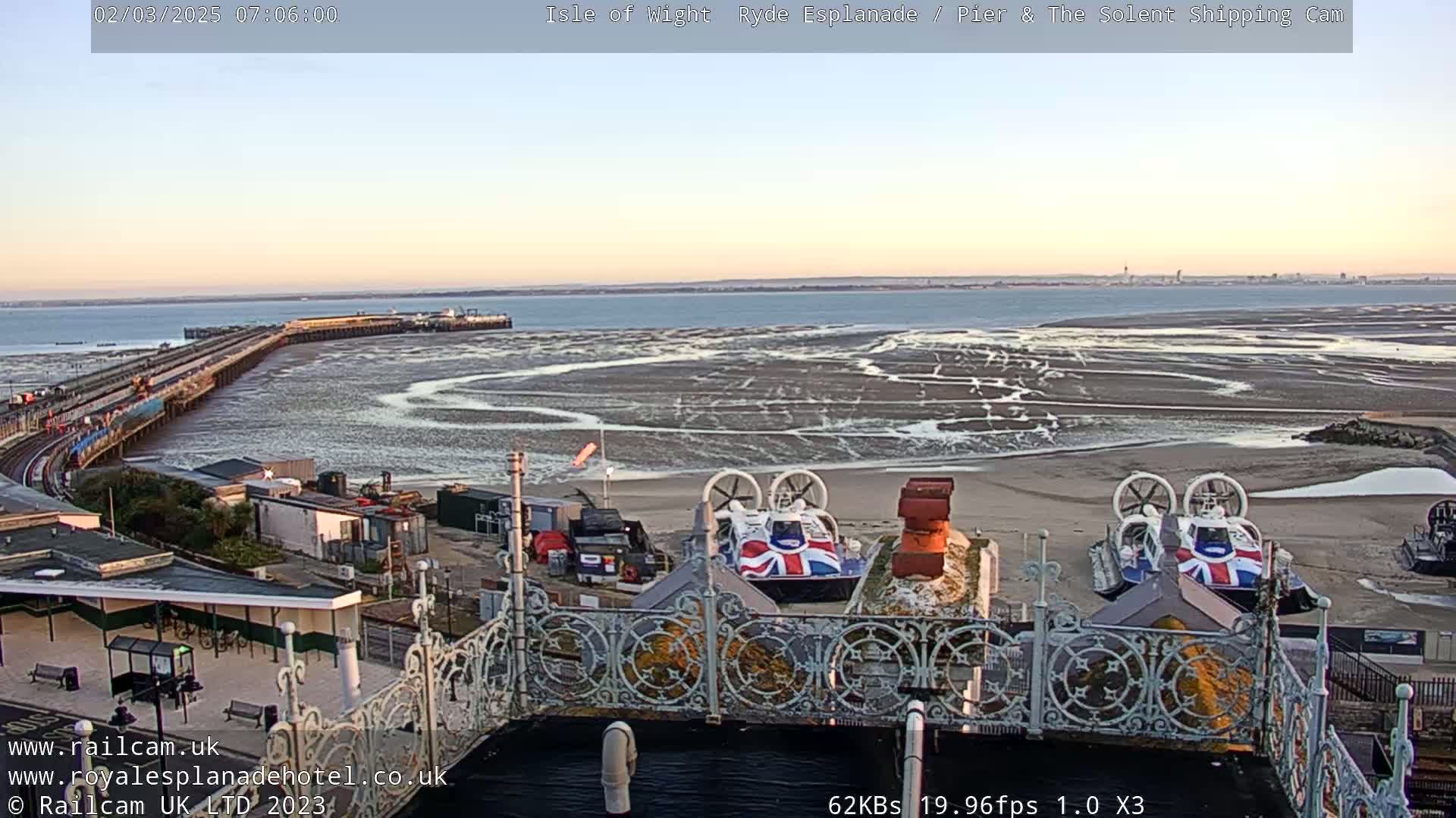 Two hovercrafts are parked on a sandy shore near a pier under a clear sunrise sky.