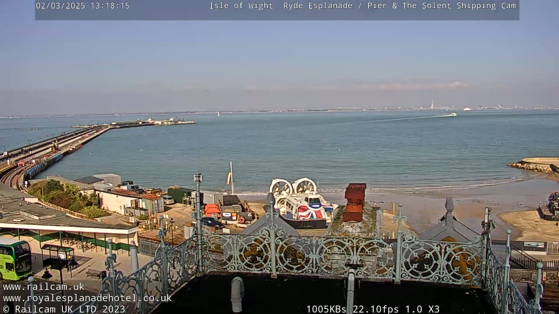 A hovercraft sits on a sandy beach near a pier extending into a calm, sunny sea.
