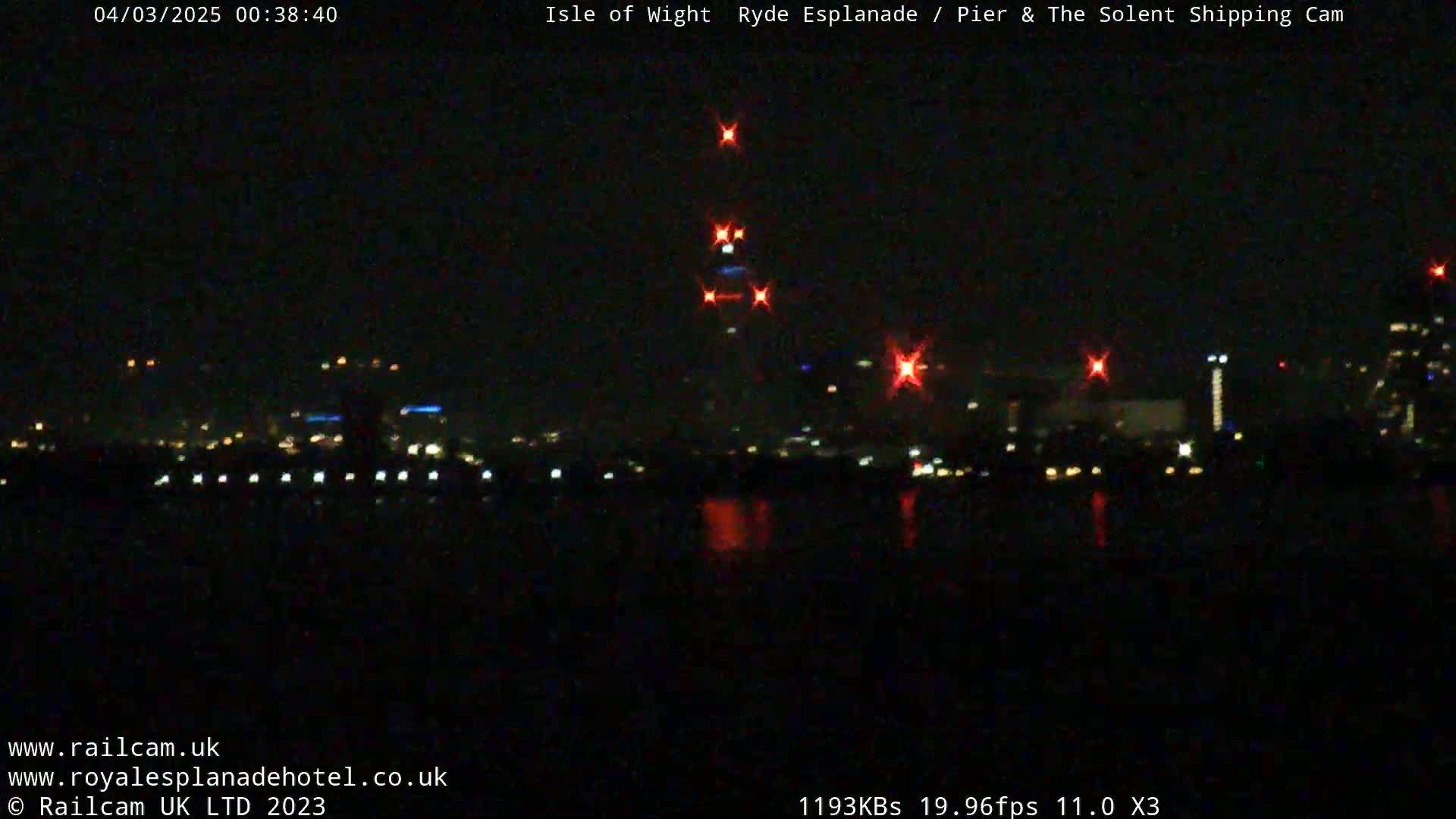 A dark nighttime view of a city skyline across a body of water, with several bright red lights visible.