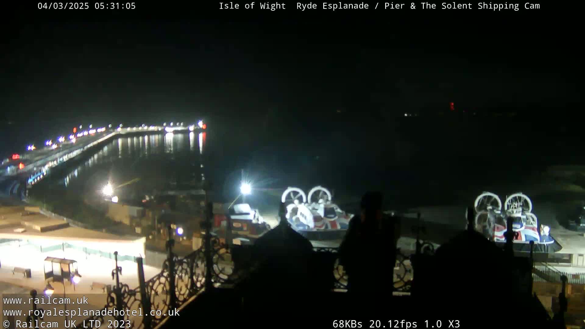 A nighttime view of a harbor with a hovercraft docked, and a brightly lit esplanade visible across the water.
