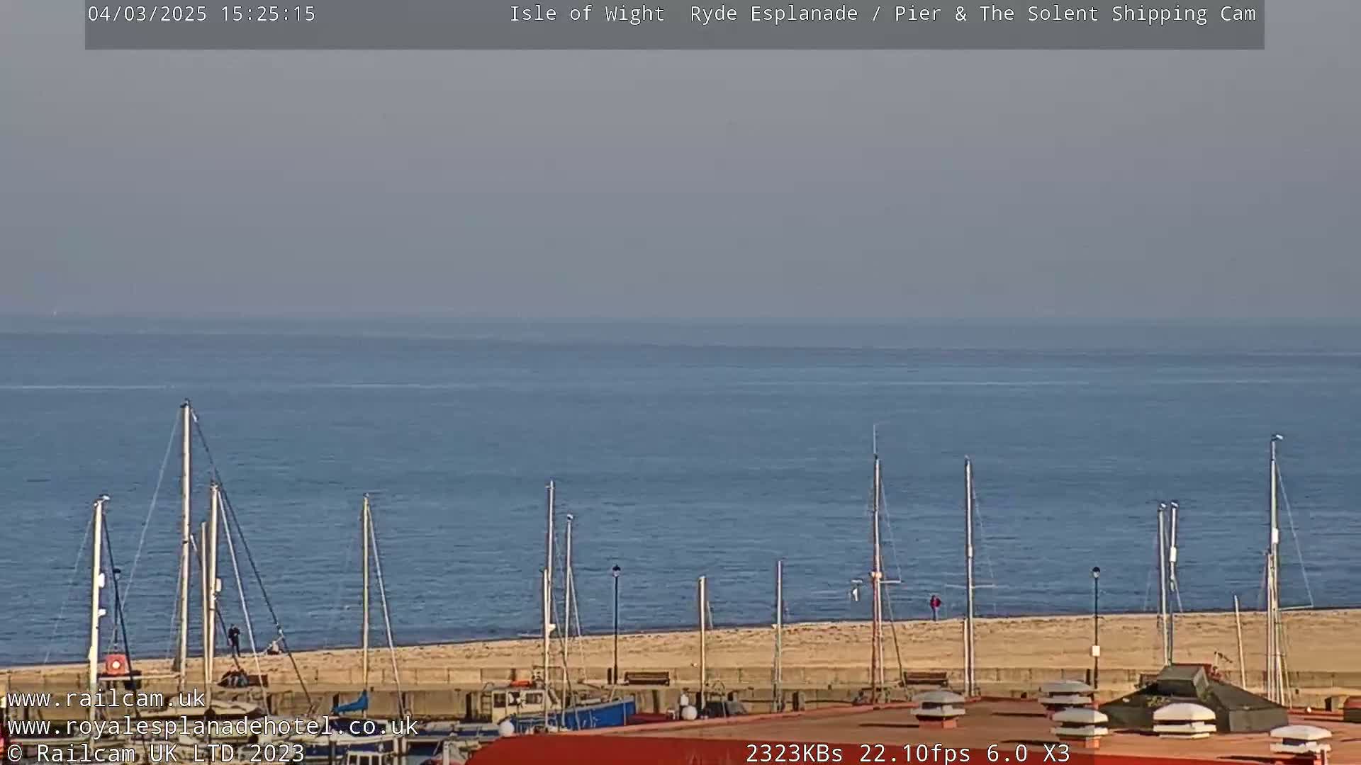 A calm, hazy day overlooks a sandy beach with several sailboats moored near a waterfront building.