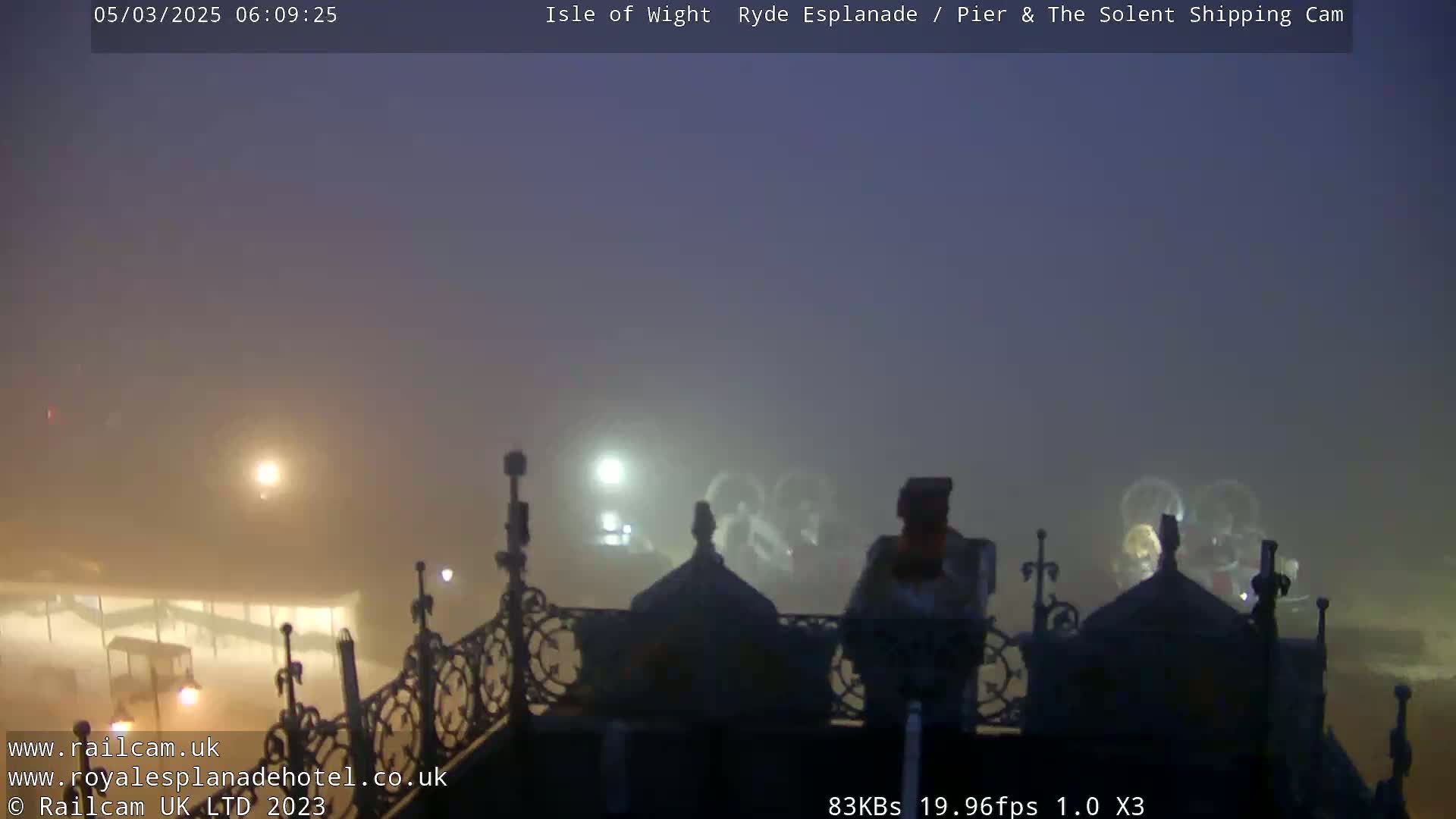A foggy, pre-dawn view from an elevated vantage point shows an ornate railing overlooking a dimly lit waterfront.