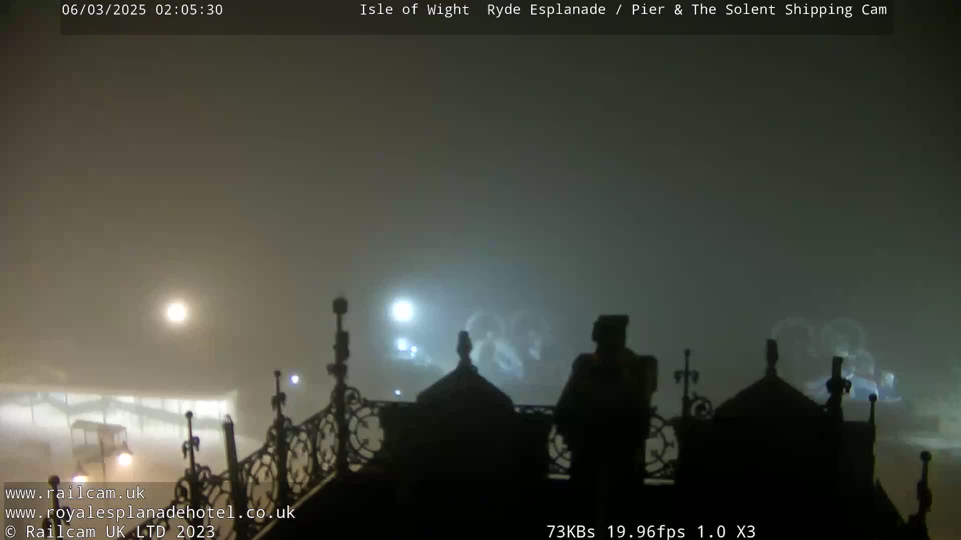 A foggy nighttime scene shows the silhouettes of buildings and ornate railings, with faint lights visible in the distance.