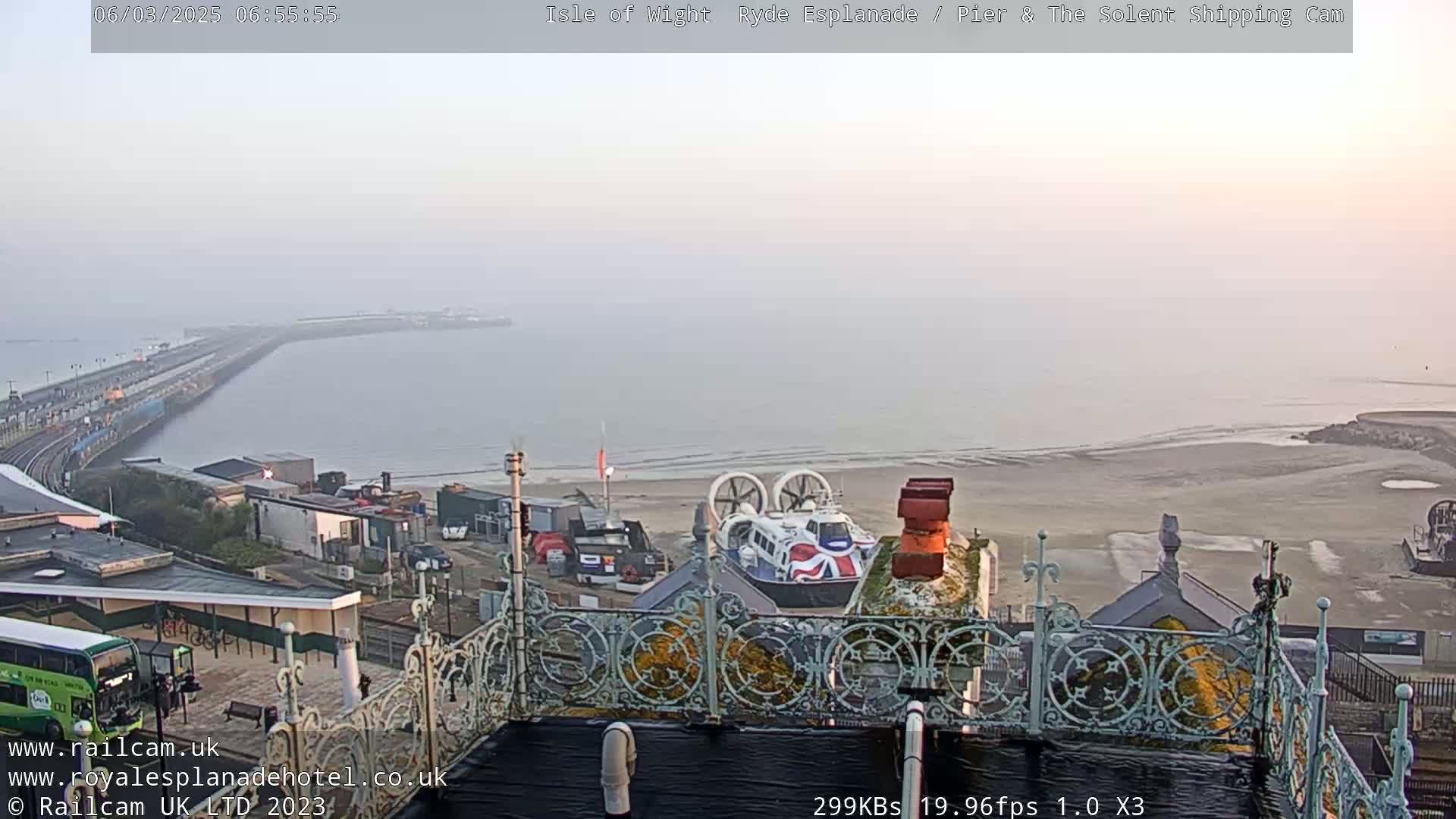 A hazy, overcast morning view from an elevated position shows a hovercraft on a sandy beach, a pier extending into the sea, and a coastal town with buildings and a bus.