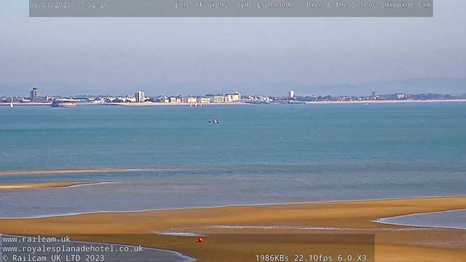 A long shot shows a sandy beach and calm, teal-colored water with a distant cityscape under a clear sky.