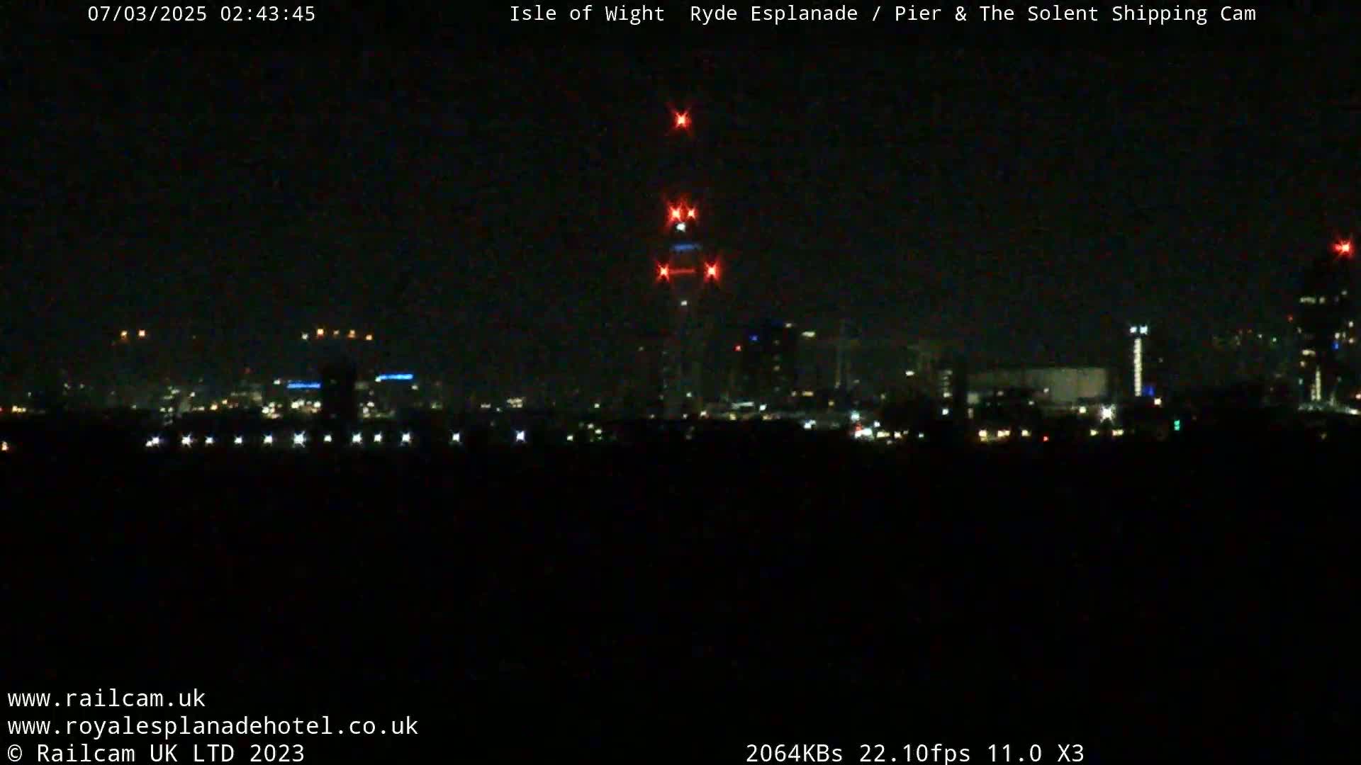 A nighttime long shot of a city skyline with a tall structure exhibiting red lights at its top, under a dark, clear sky.