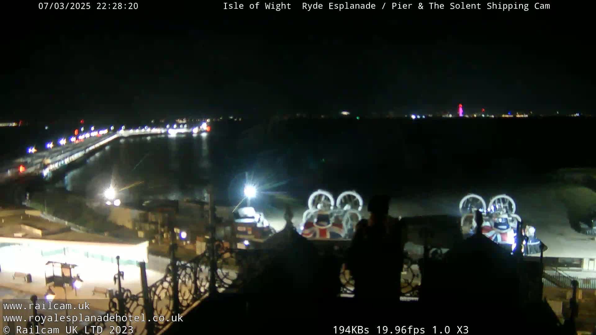 A nighttime, overhead view of a pier and esplanade with a body of water, featuring several illuminated vehicles and what appears to be a hovercraft.
