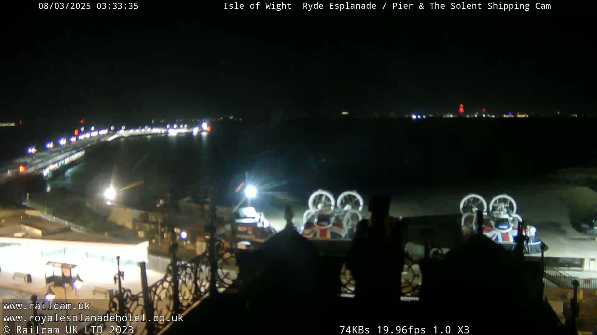 A nighttime, long shot of a pier and body of water with a hovercraft and some lights visible in the distance.