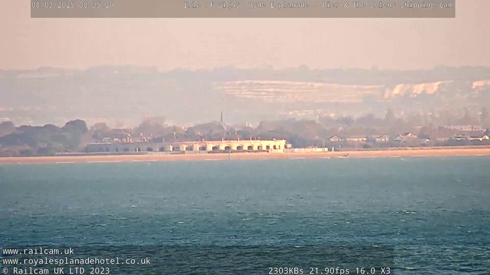 A hazy, long shot shows a building on a sandy beach, with calm water in the foreground and distant hills under a light-colored sky.