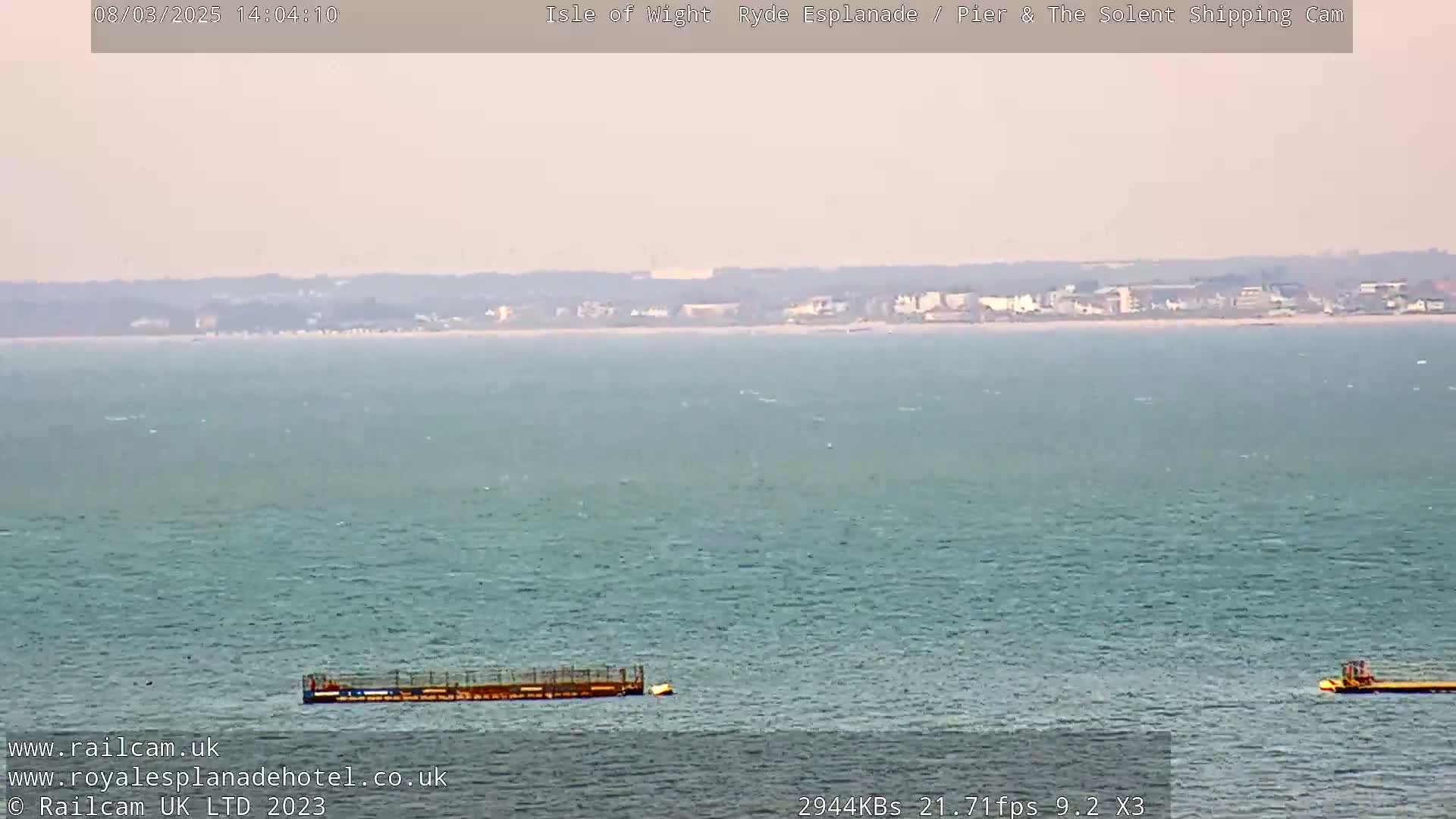 A hazy, calm sea with a distant shoreline and two small structures floating on the water.