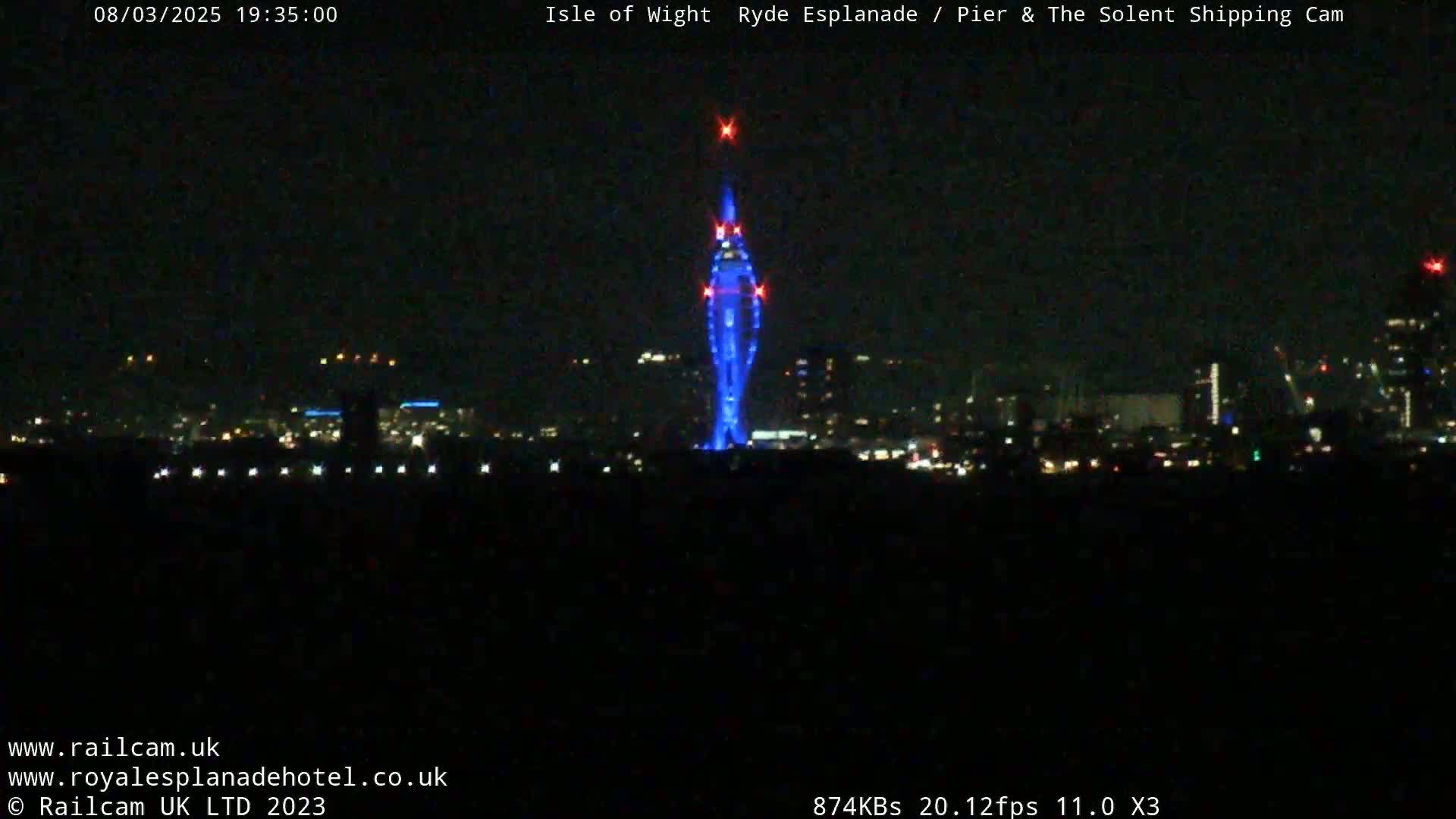 A tall, slender structure illuminated in blue is seen at night against a dark city skyline.
