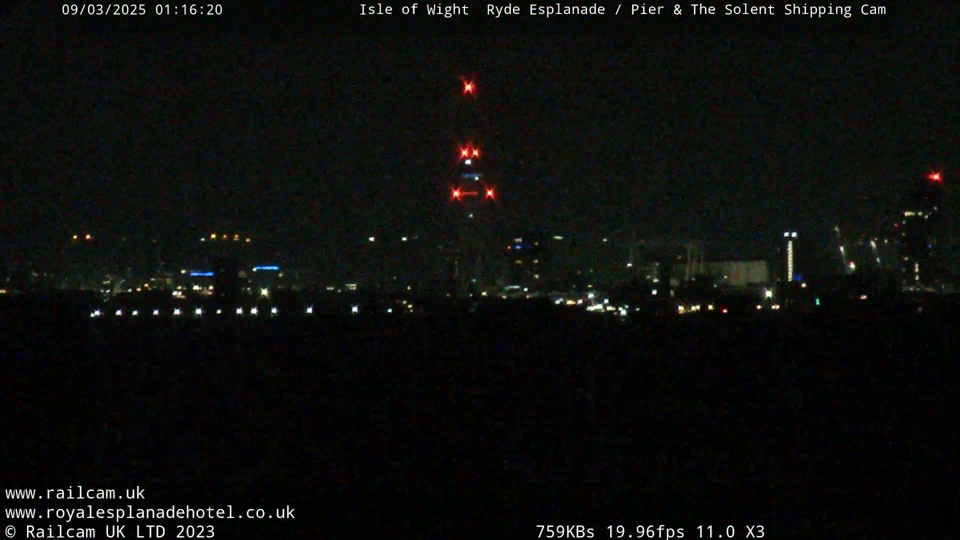 A nighttime long shot shows a distant city skyline illuminated with lights, a tall structure with red lights atop it, under a clear, dark sky.