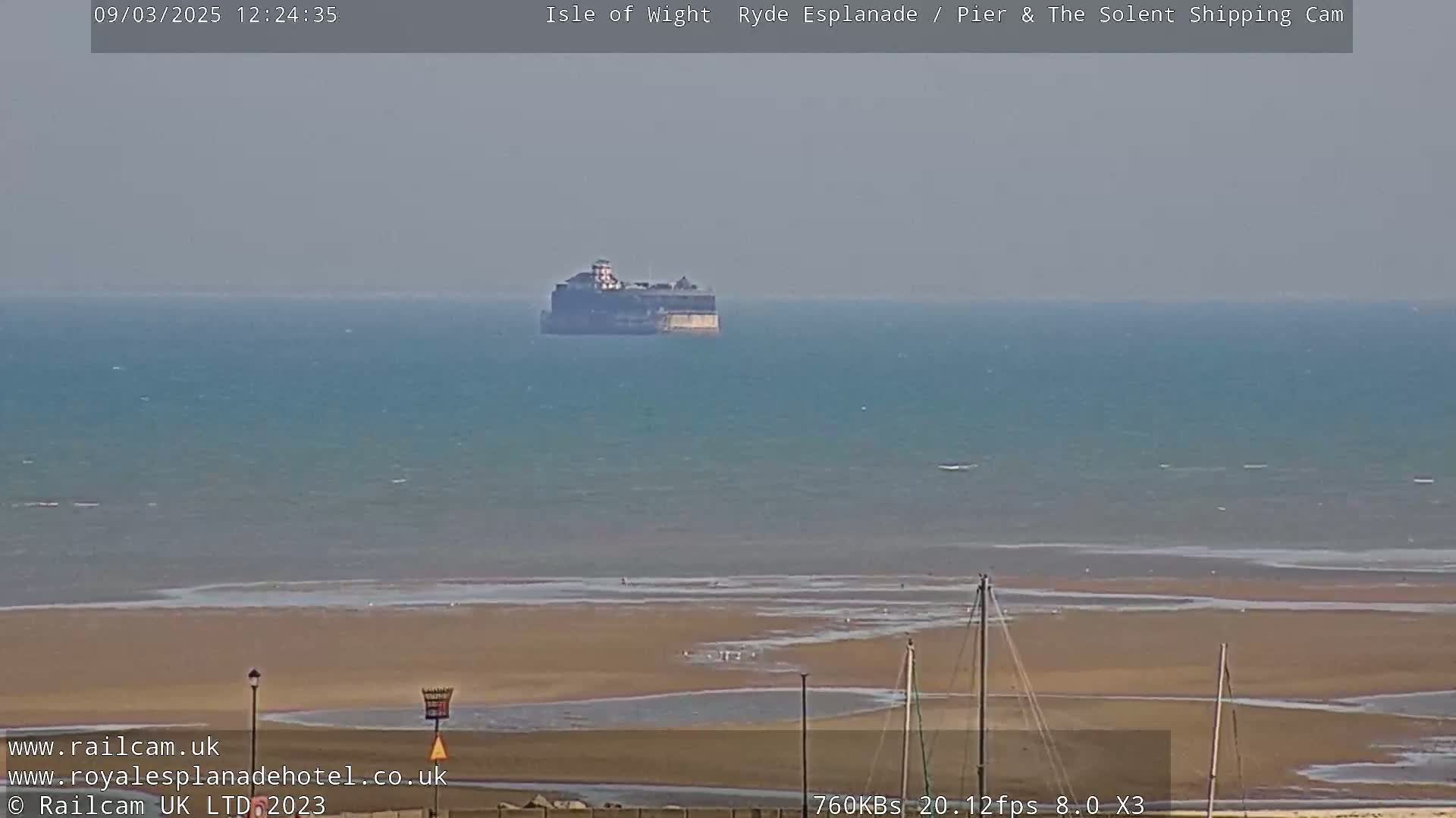 A hazy, distant view of a large structure in the sea, a sandy beach, and several masts on the shore.