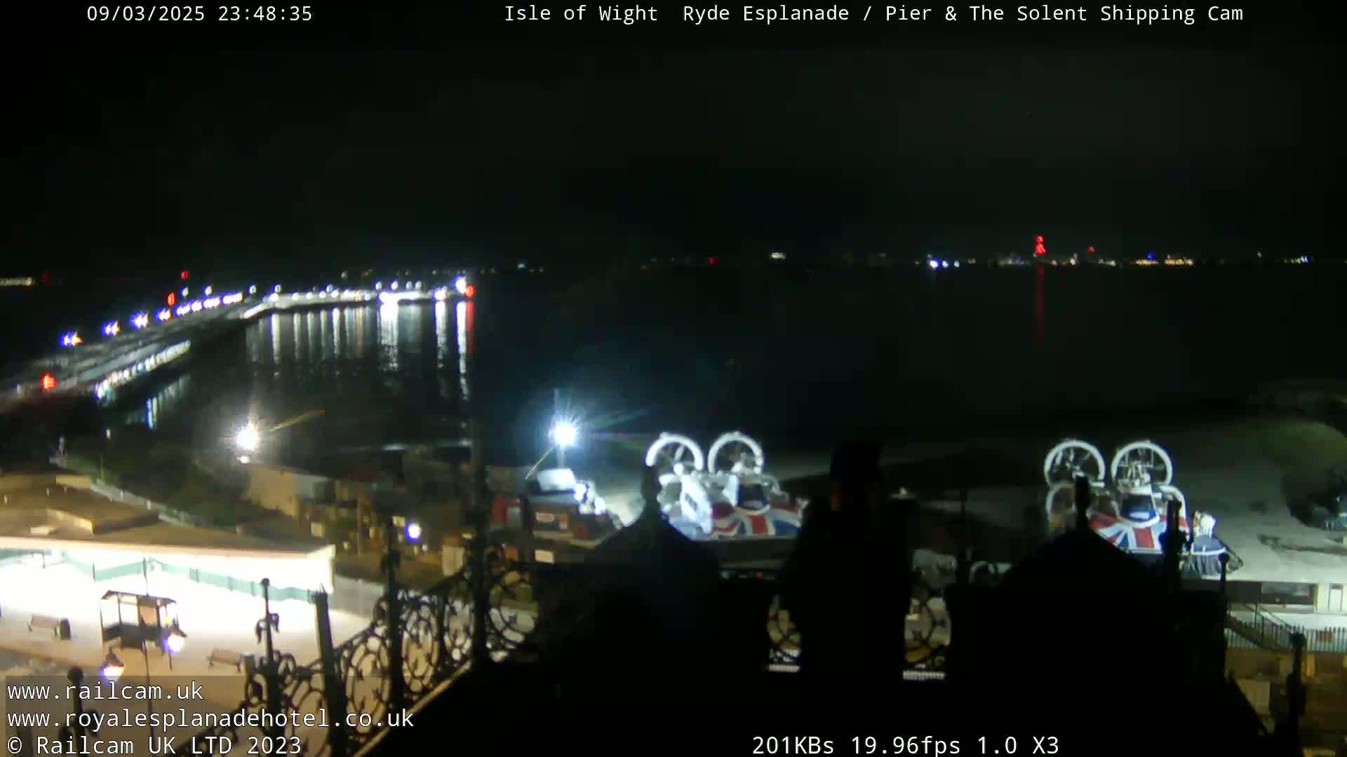 A nighttime view of a pier and body of water, with two hovercrafts parked on the shore and lights illuminating the scene.