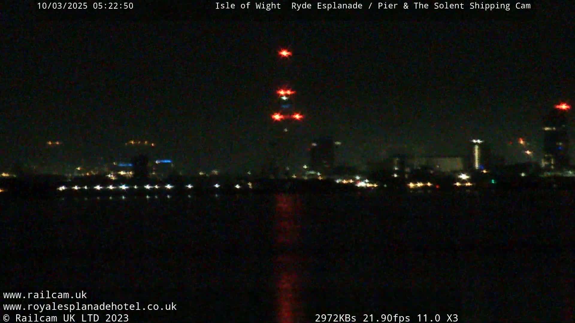 A nighttime view across dark water shows a distant cityscape with scattered lights, punctuated by a series of bright red lights in a vertical line.
