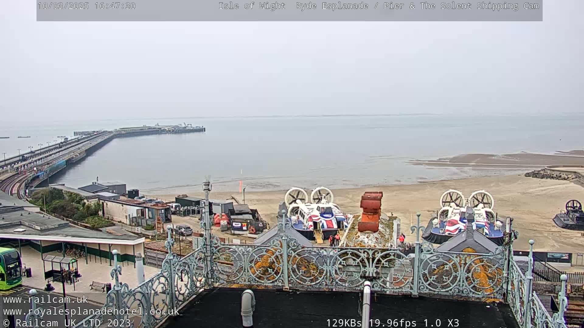 A view from a high vantage point shows a long pier extending into a calm, gray sea under an overcast sky, with a hovercraft resting on a sandy beach in the foreground.