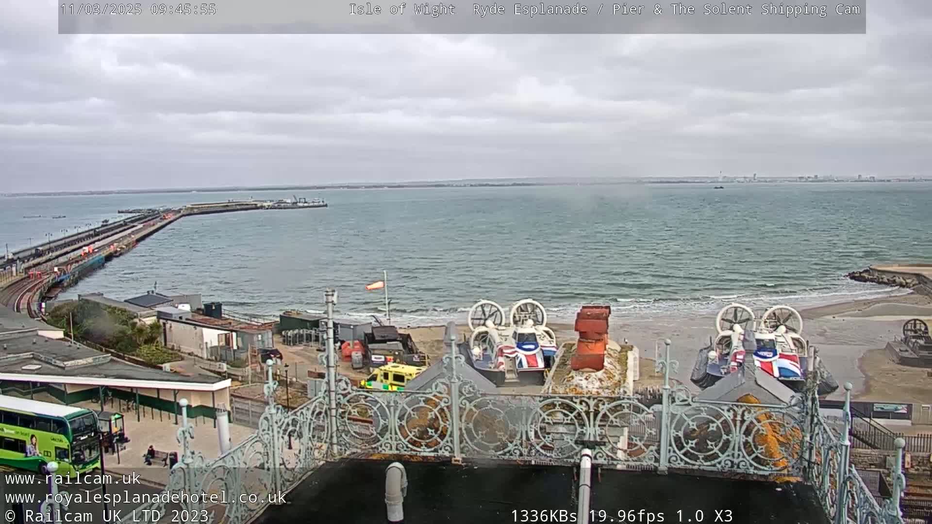 Overcast skies loom over a coastal scene featuring a pier extending into the sea, two hovercrafts on a sandy beach, and a bus at a station.
