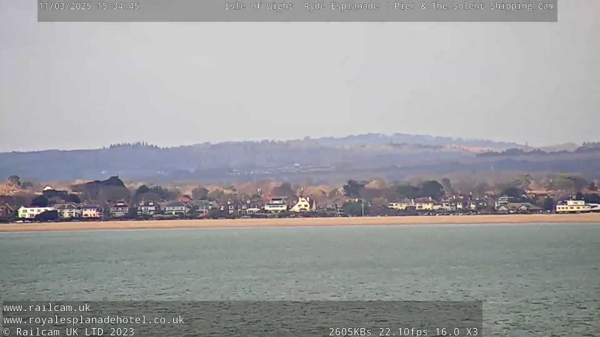 A hazy view shows a calm body of water with a sandy beach and a line of buildings nestled against hills in the background.