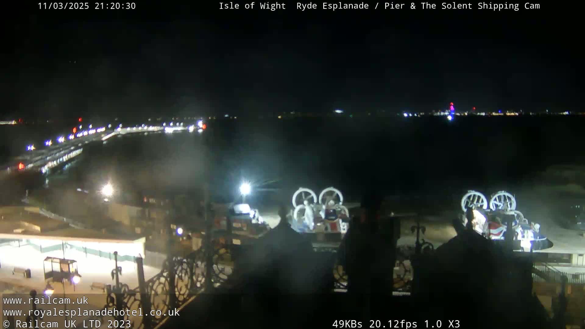 A nighttime view of a body of water with lights across the horizon, two hovercrafts on the shore, and a pier in the foreground.