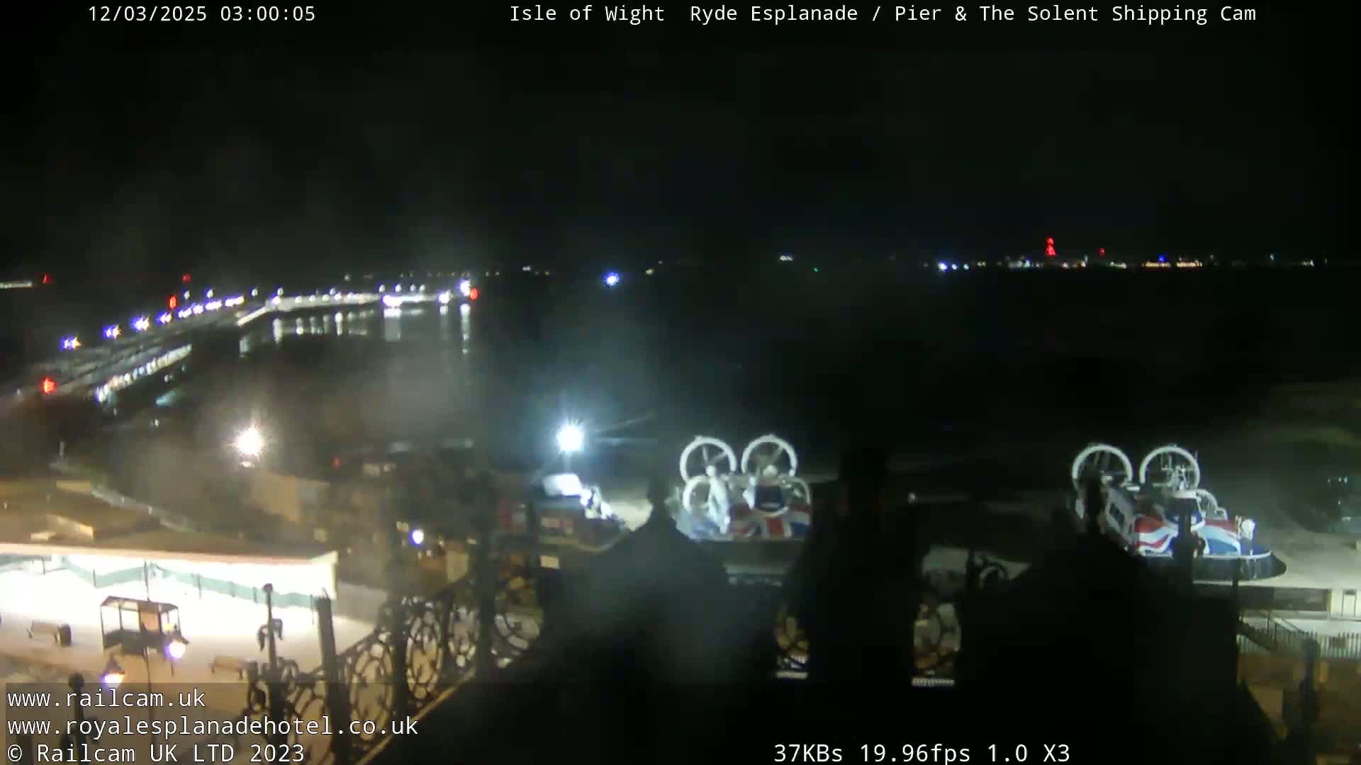 At night, two hovercrafts are parked near a body of water with lights visible along a shoreline in the distance.