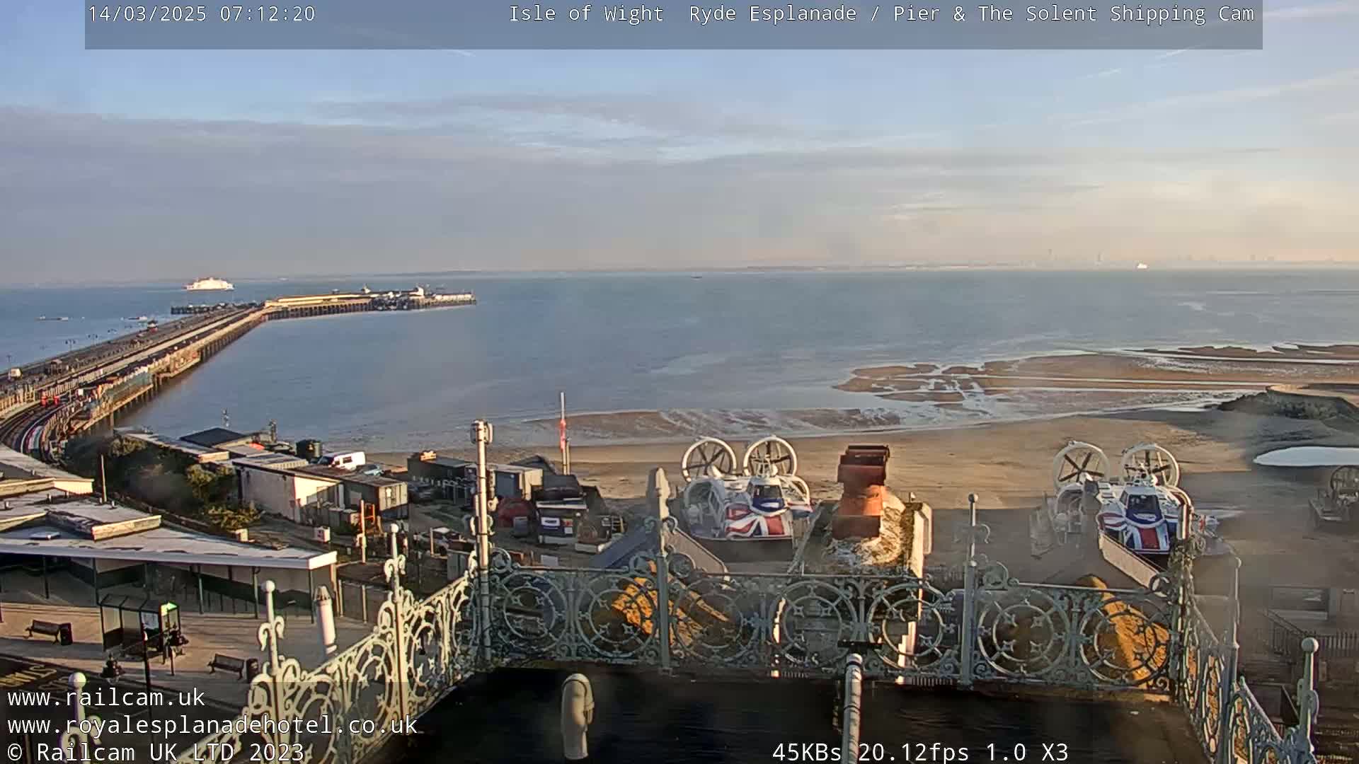 A long pier extends into a calm body of water under a mostly clear sky, with two hovercrafts parked on a sandy beach in the foreground.