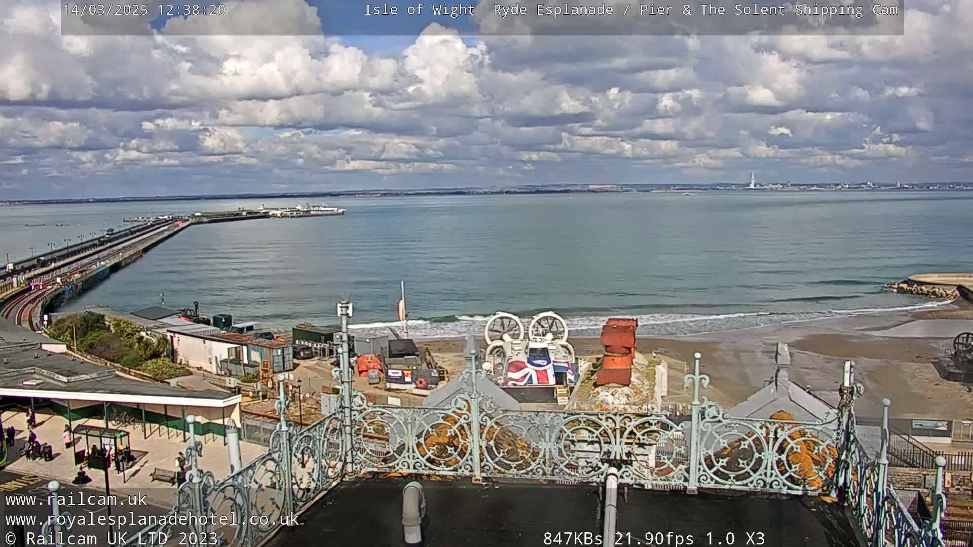 A partly cloudy day shows a long pier extending into a calm sea with a sandy beach and a hovercraft on shore.