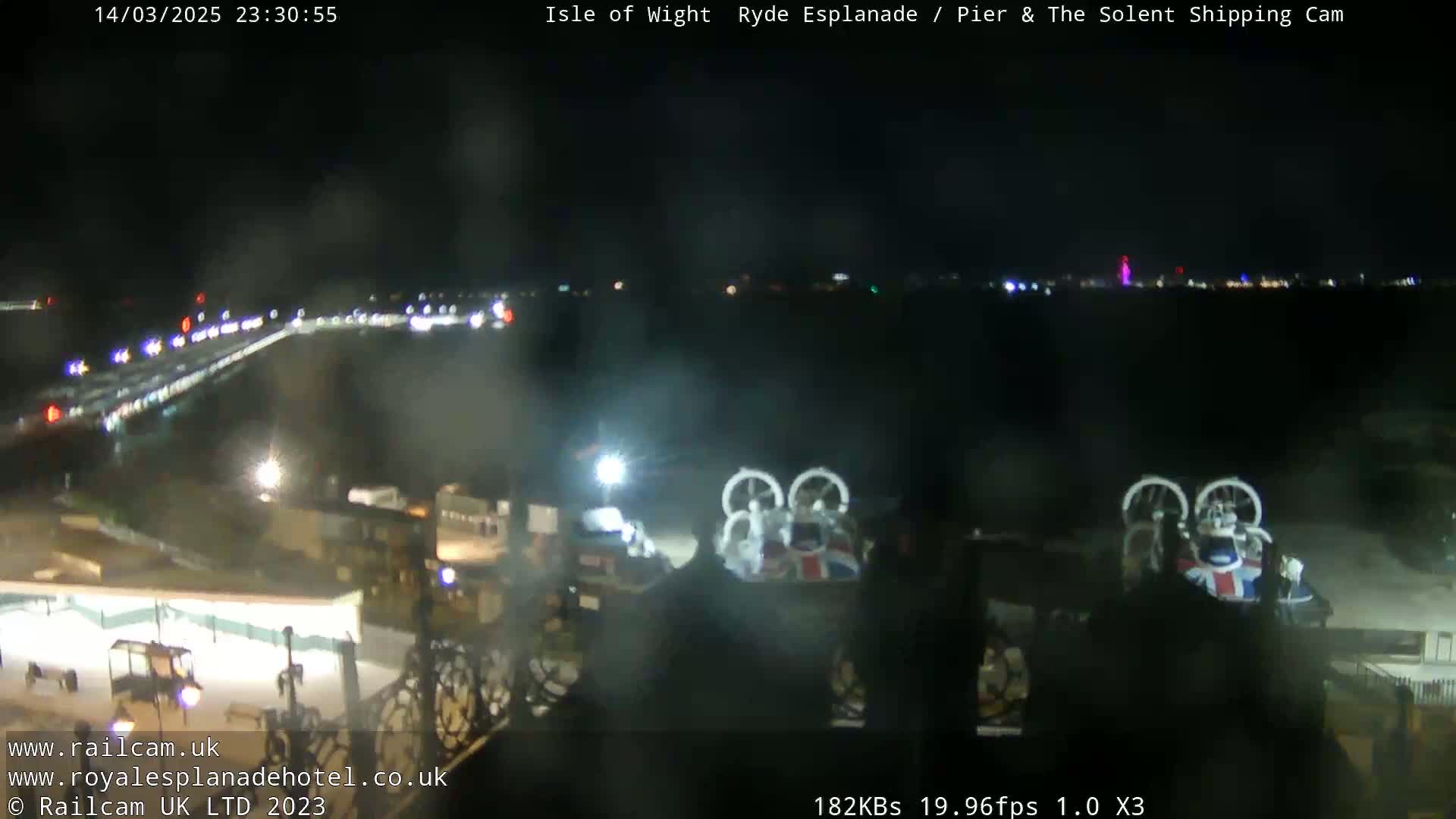 A nighttime view of a pier and waterfront area with a few vehicles visible, and a hovercraft docked nearby under hazy conditions.